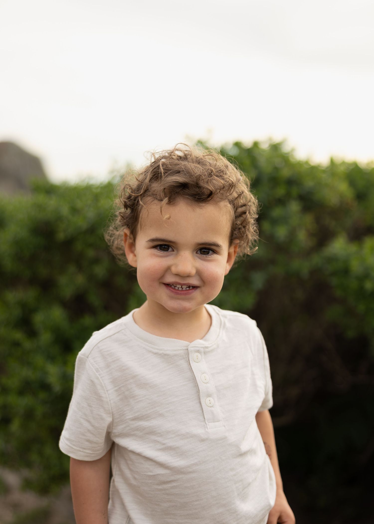 Smiling young boy with curly hair wearing a white henley shirt outdoors with green bushes behind him.