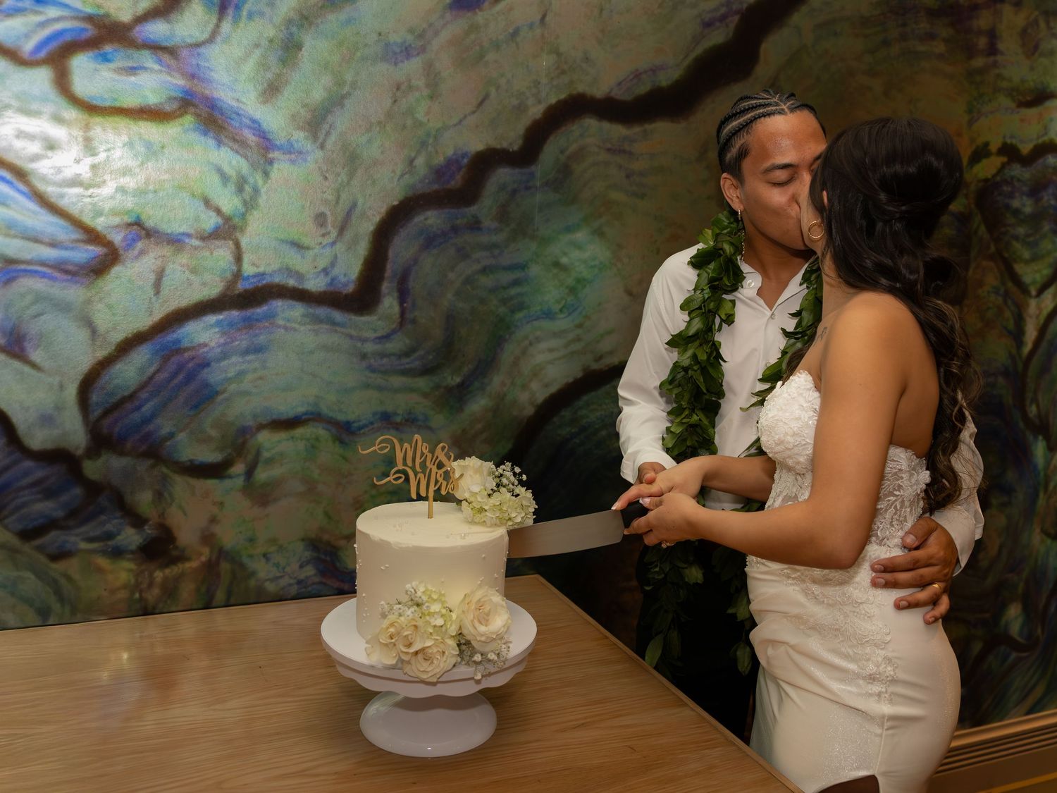 Newlywed couple cutting white wedding cake, groom wearing green lei, elegant marble wall backdrop.