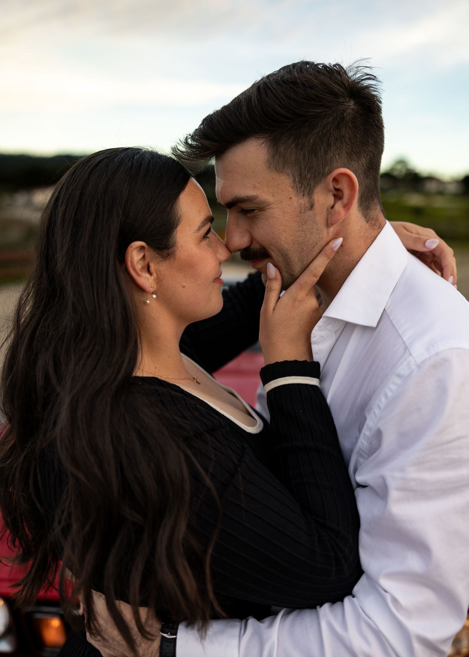 A romantic couple sharing an intimate moment, nearly kissing outdoors at dusk, woman in black top, man in white shirt.