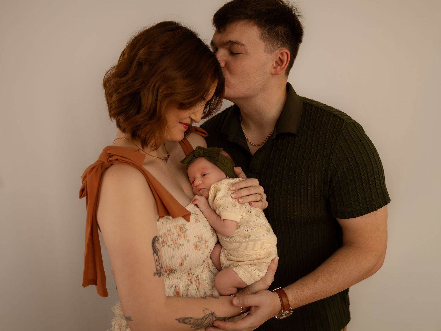 A couple tenderly holds their newborn baby, with the man kissing the womans head against a soft white background.