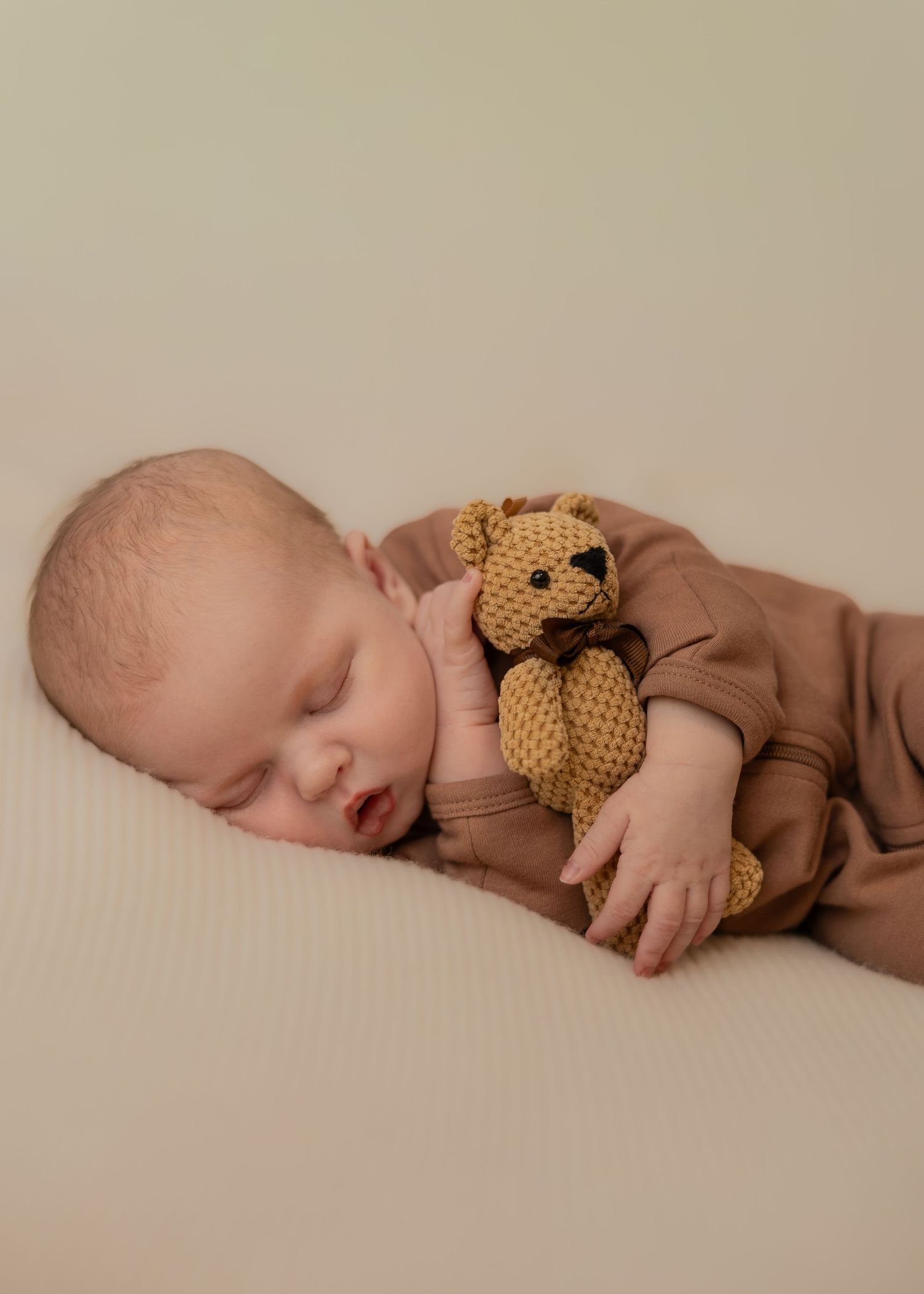 Sleeping newborn baby in brown romper cuddling a small knitted teddy bear on a cream background.