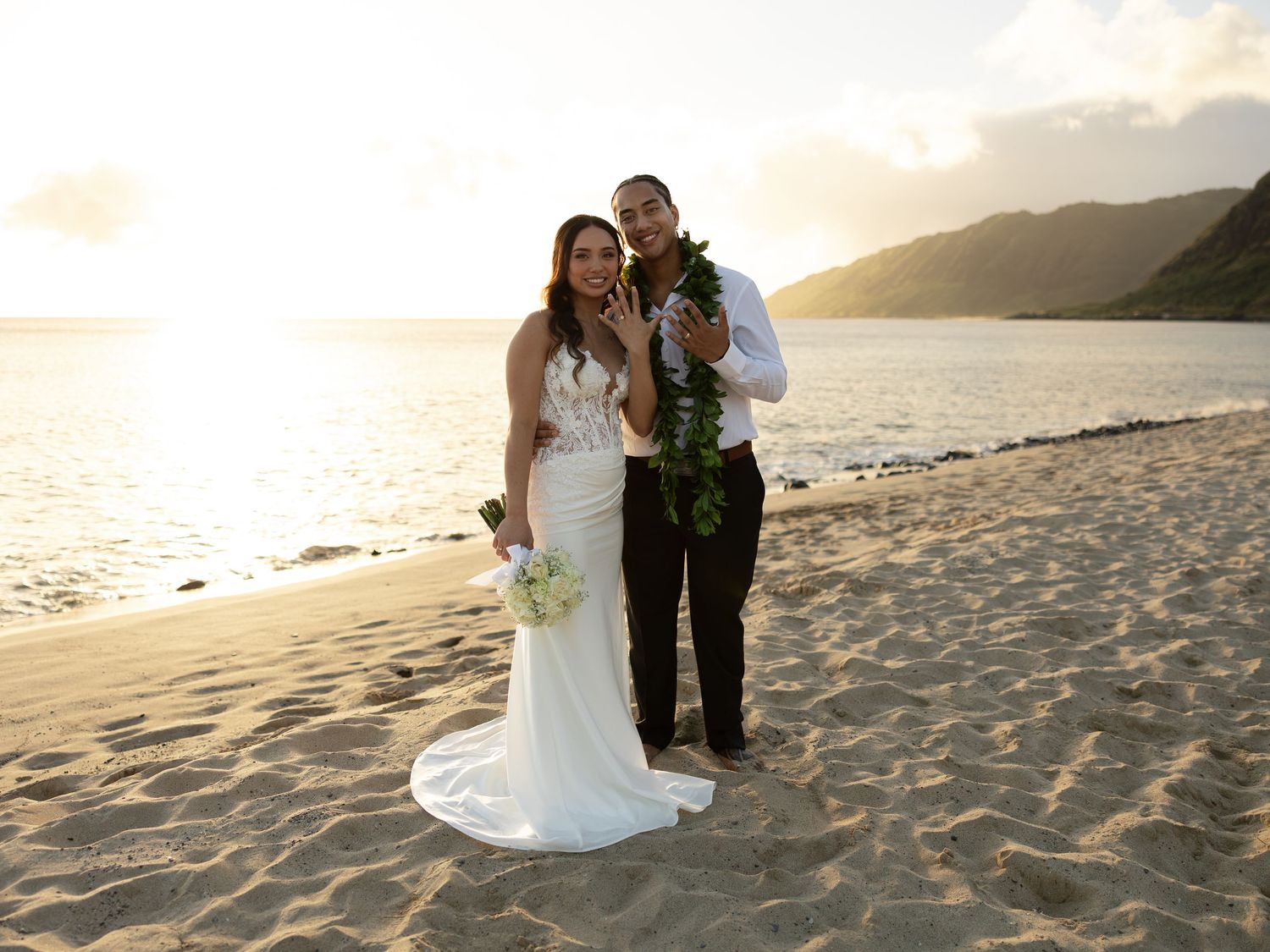 Newlywed couple posing on a sandy beach at sunset, bride in white gown holding bouquet, groom wearing floral lei.