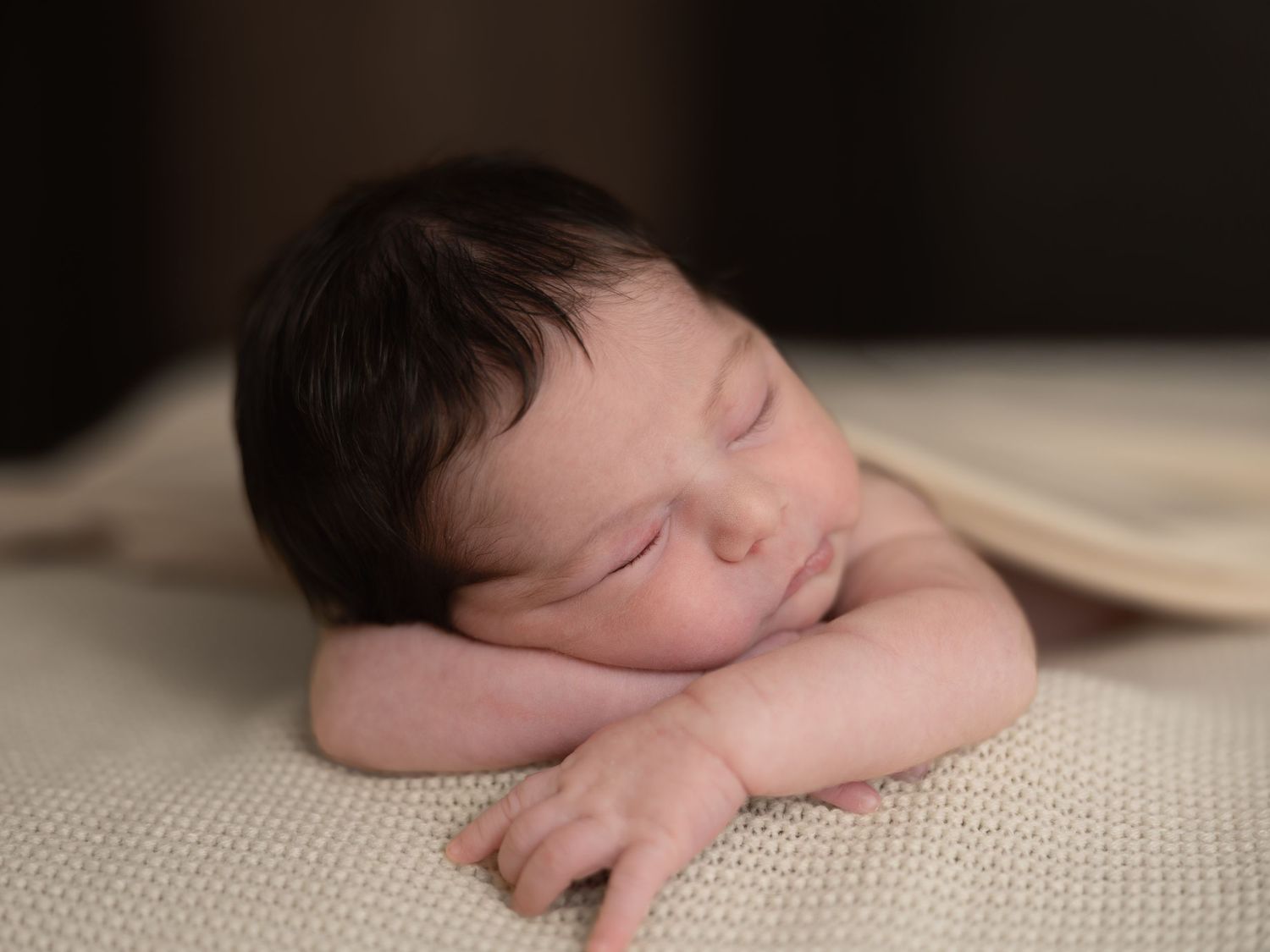 Peaceful newborn baby sleeping on a soft cream knit blanket with dark hair and tiny folded hands.
