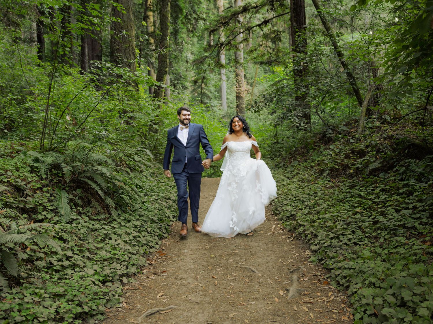Bride and groom walking hand-in-hand on a lush forest trail surrounded by green ferns and towering trees.