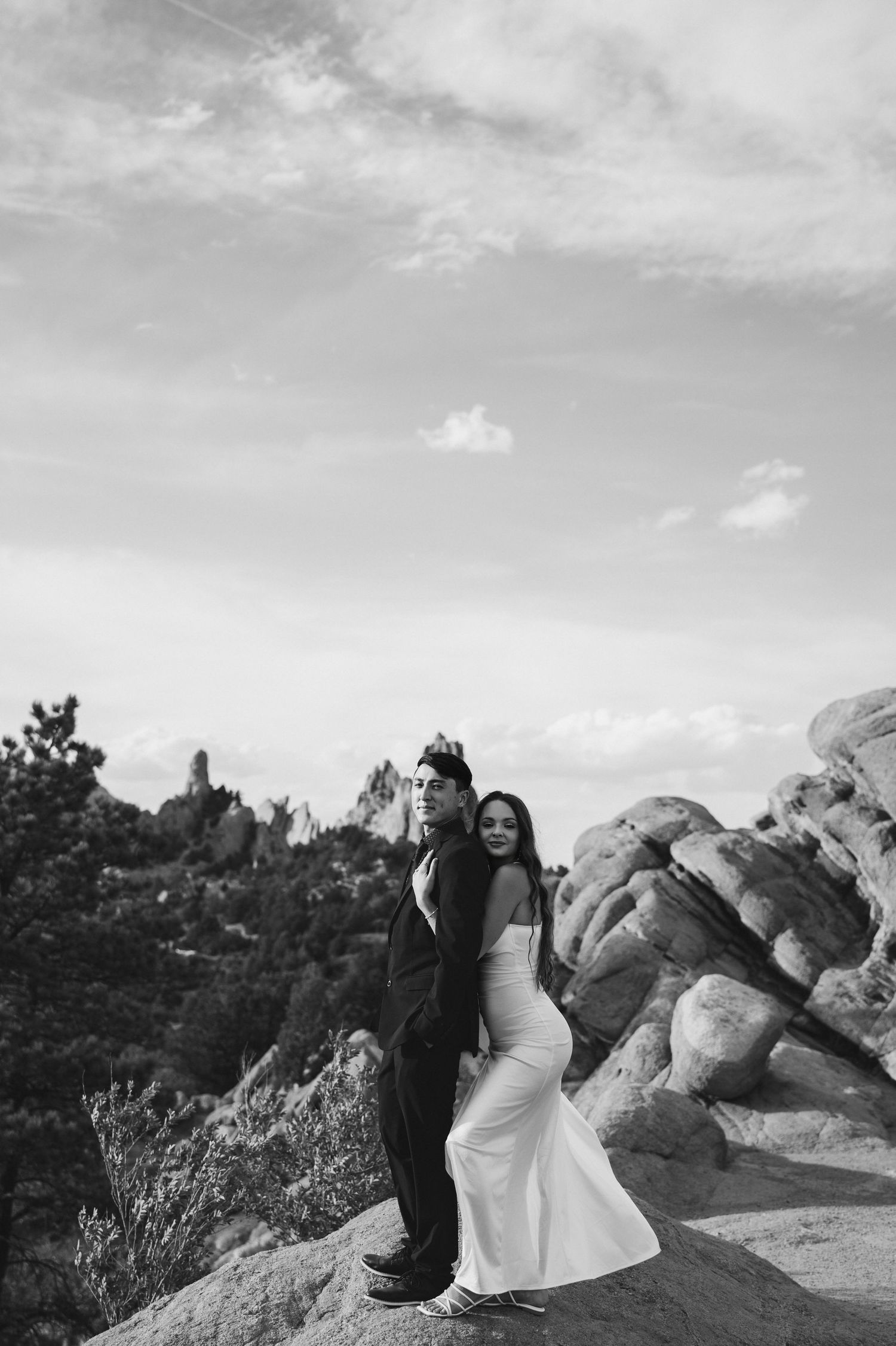A black and white photograph captures a joyful couple posing against rocky desert terrain and cloudy skies.