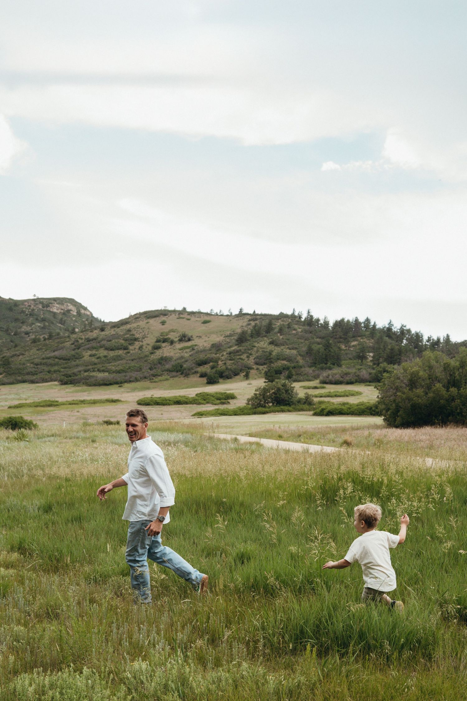 Two people run and play together across a scenic green meadow with rolling hills and trees in the background.