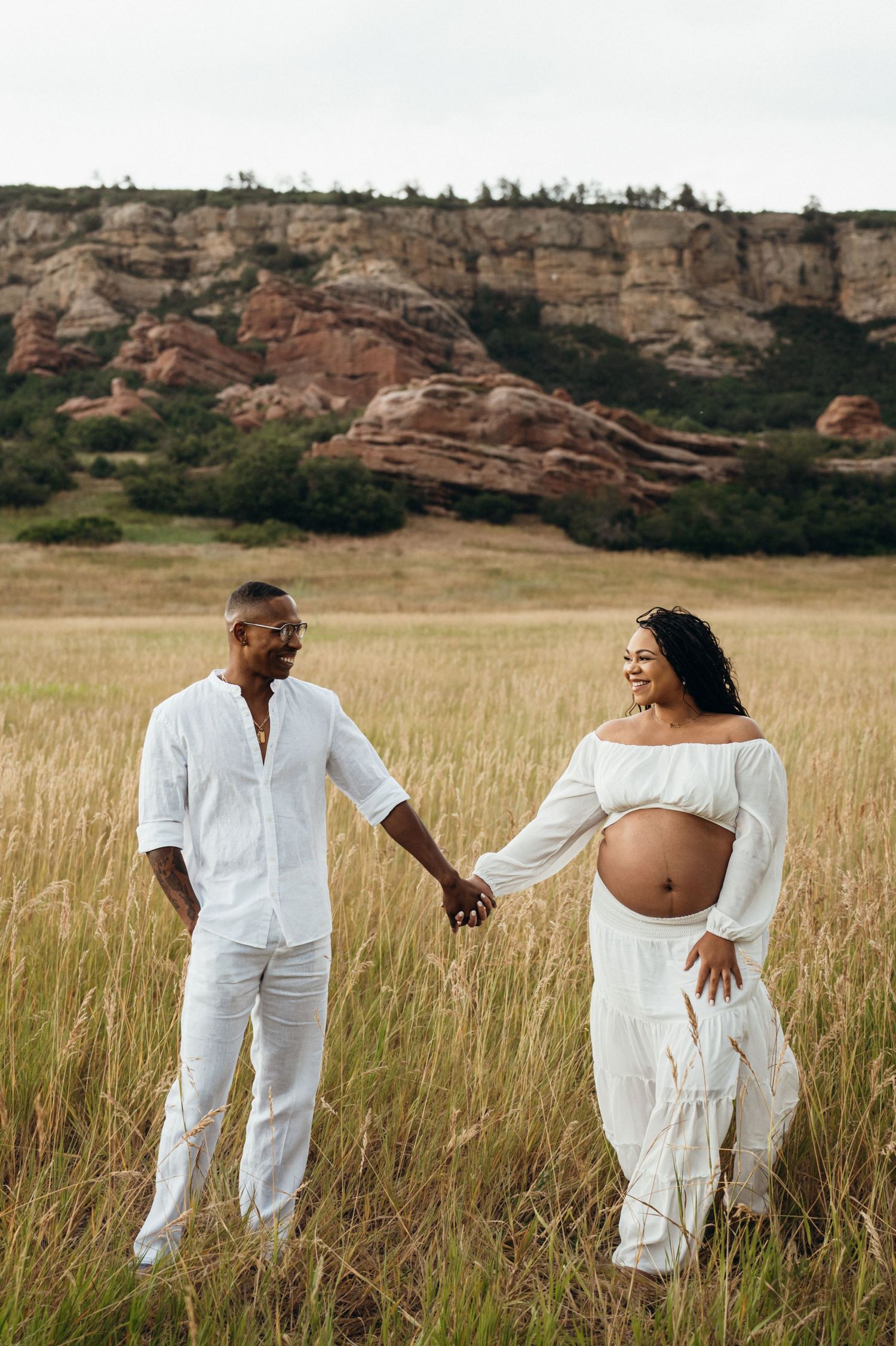 A couple in white outfits holding hands in a grassy field with dramatic red rock cliffs in the background.