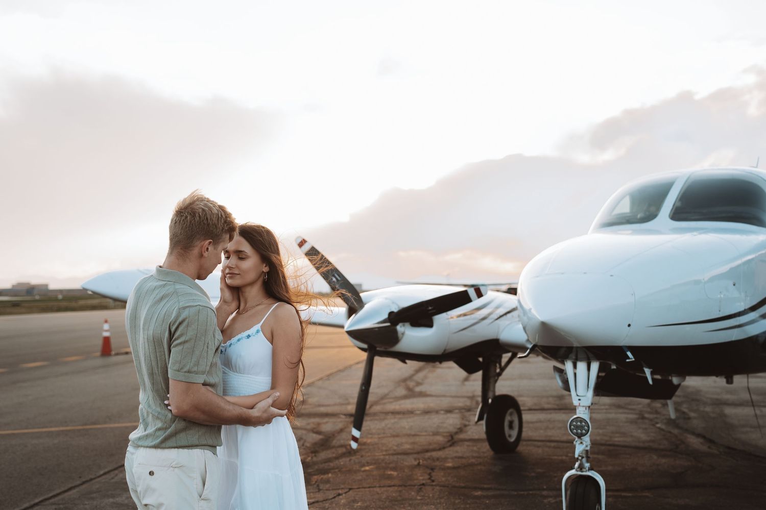 A romantic couple embraces on a private airport tarmac near a small aircraft at sunset.