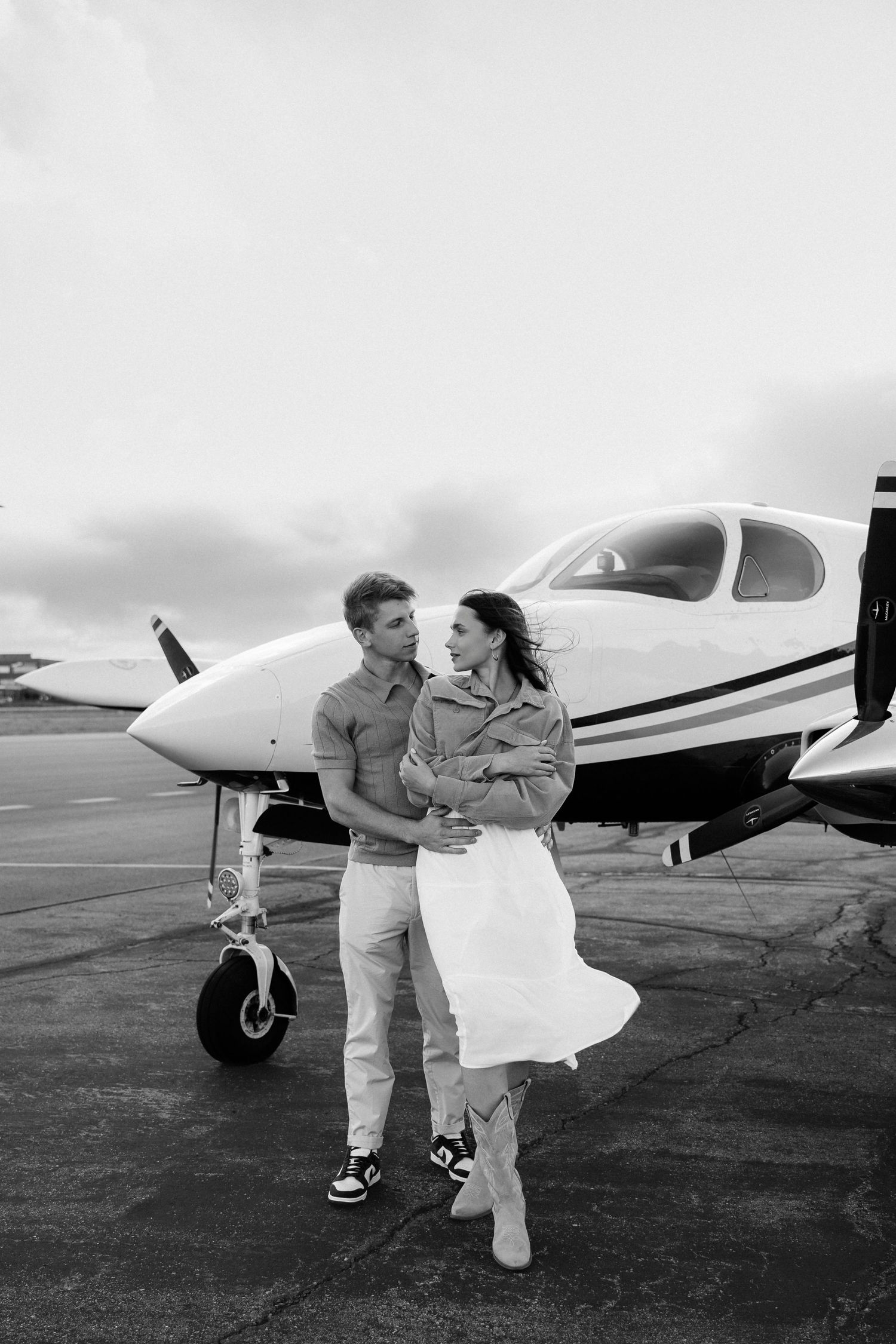 A romantic couple embrace near a small private airplane on a cloudy day at an airfield, captured in black and white.