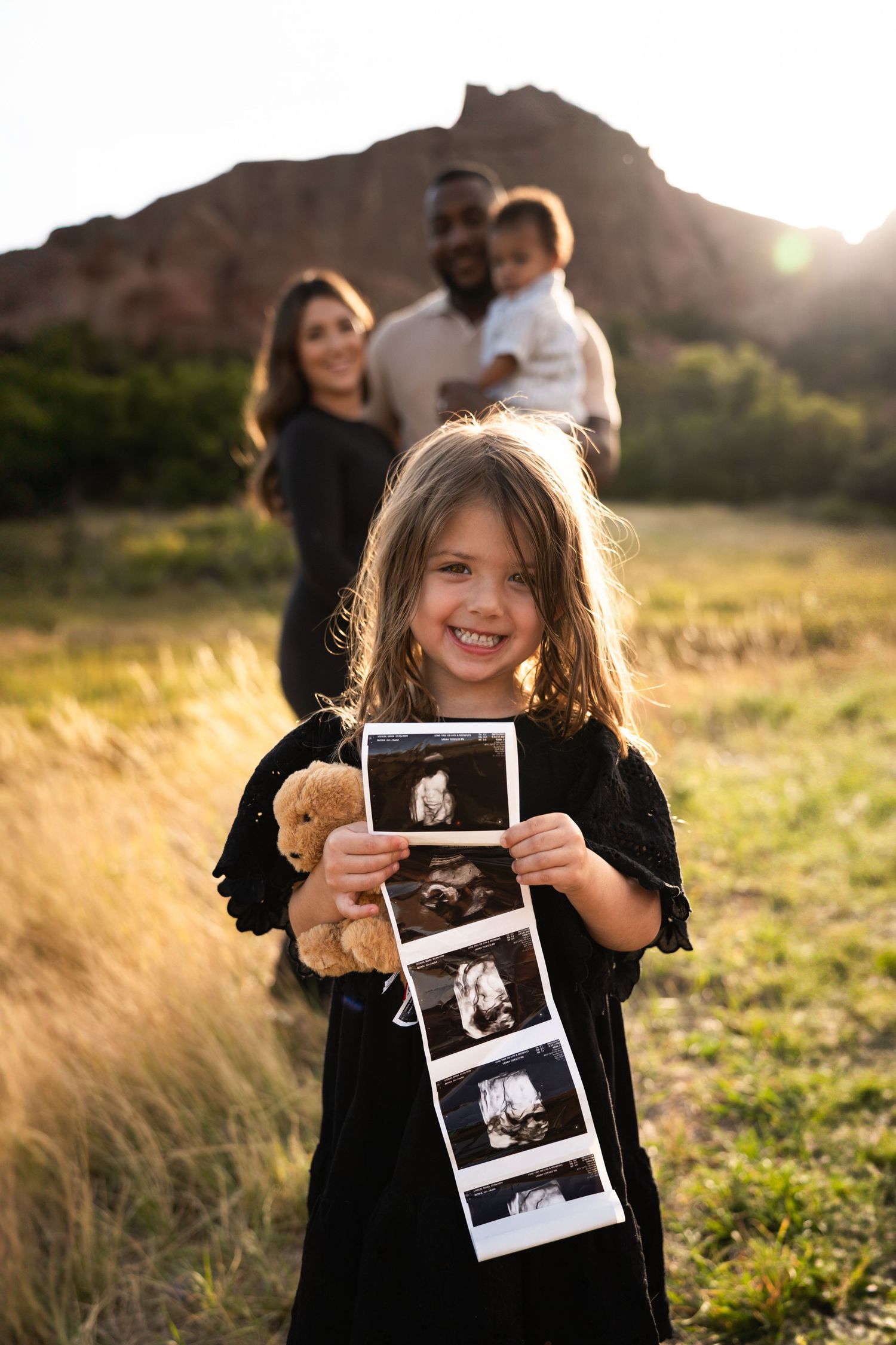 A child holds ultrasound photos while family stands in background during sunset.