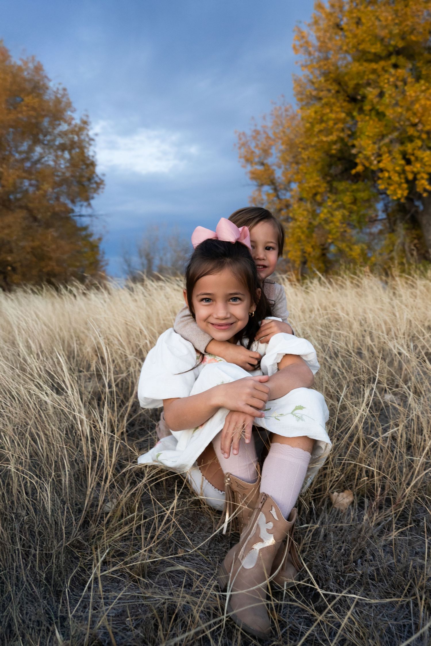 Two figures sit in golden autumn grass with yellow trees and blue sky in background.