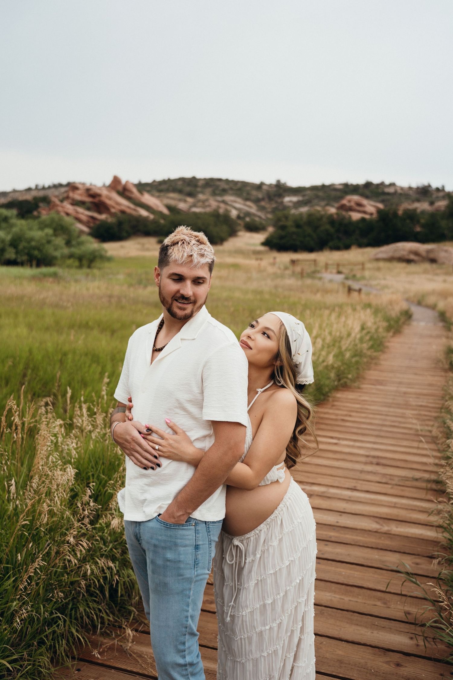 A couple stands together laughing on a wooden boardwalk path through tall grass and rolling hills.