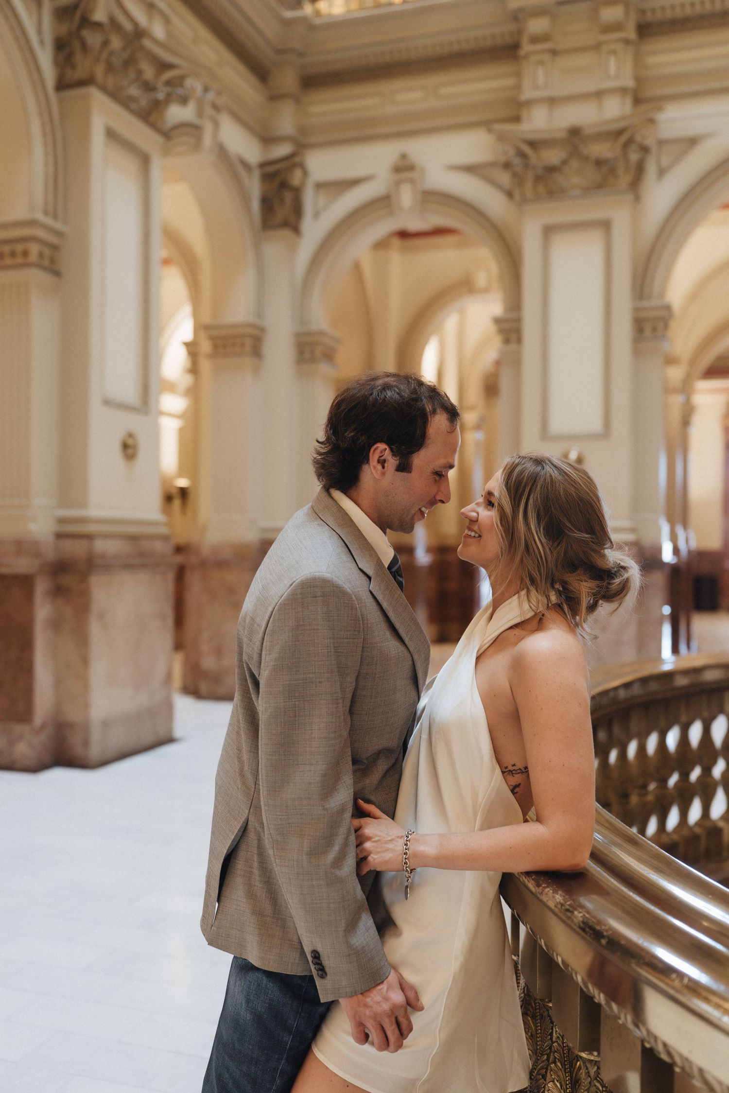 A romantic couple shares intimate moments in a grand marble hallway with ornate classical architecture and arches.