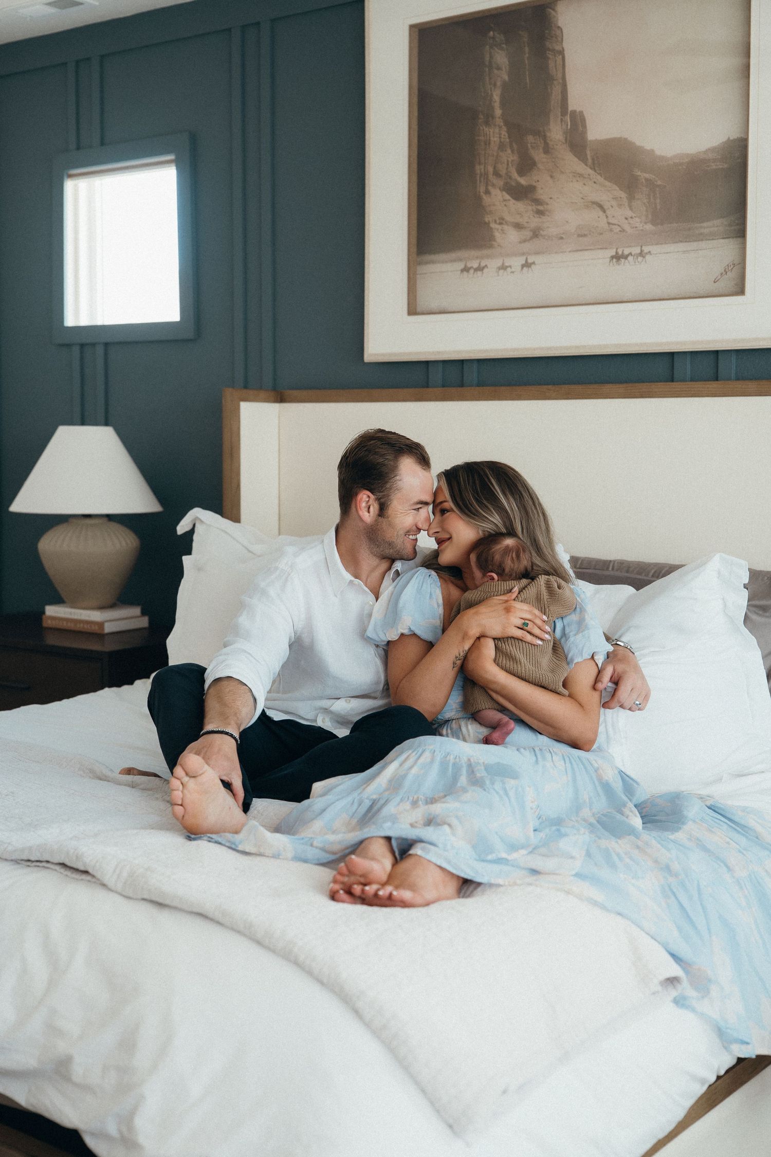 A couple shares a tender moment on a white bed holding their baby in a stylish teal bedroom with framed art.