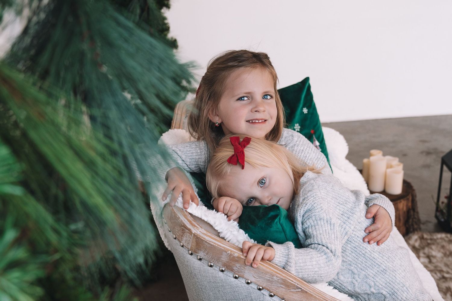 Two children in white sweaters cuddle together in a chair near a Christmas tree.