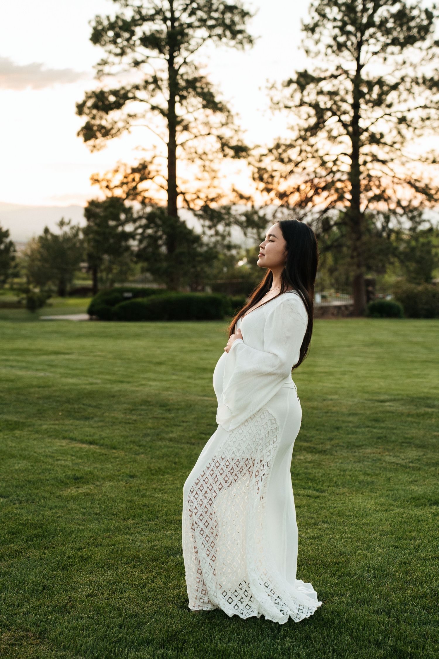 An expectant person in a flowing white lace dress poses in a grassy field at sunset with tall pine trees in the background.