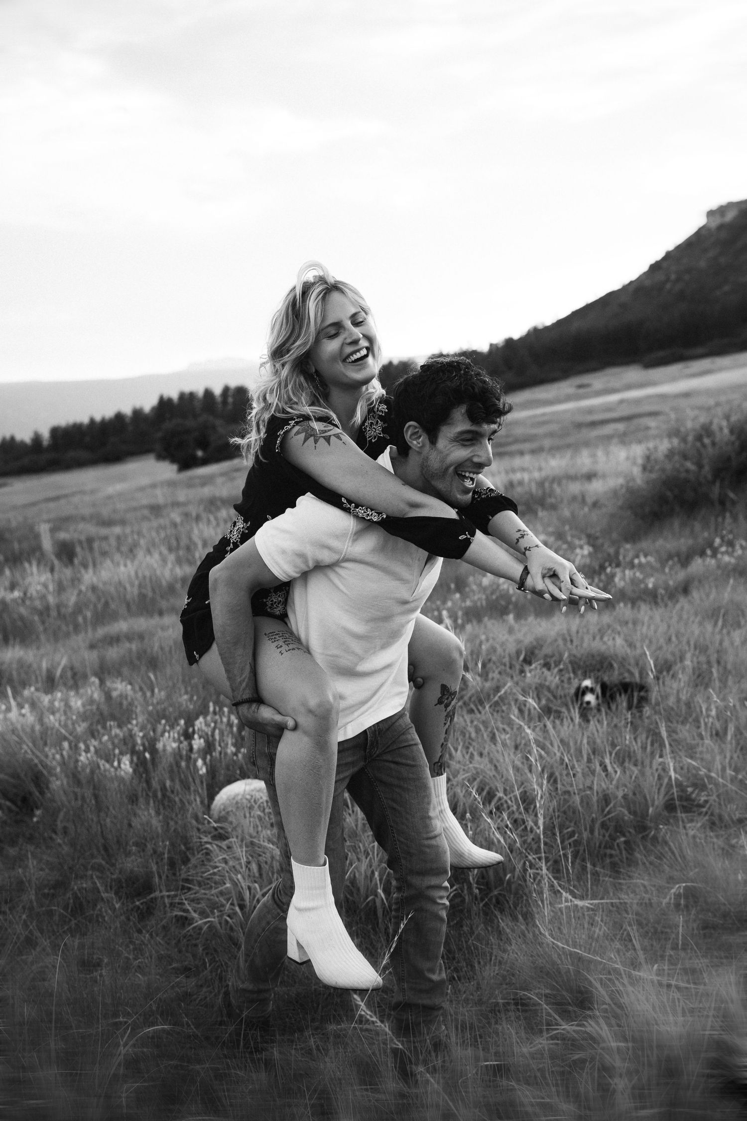 A joyful couple plays and laughs together in a scenic mountain field at sunset, captured in black and white.