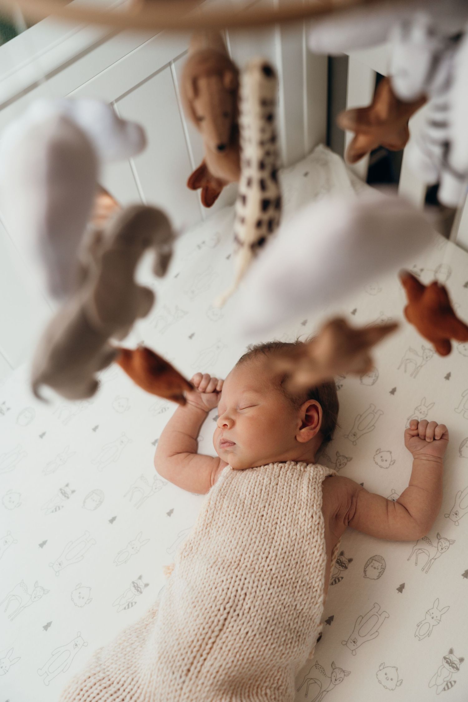 A sleeping baby wearing a cream knit romper lies in a crib beneath hanging stuffed animal toys.