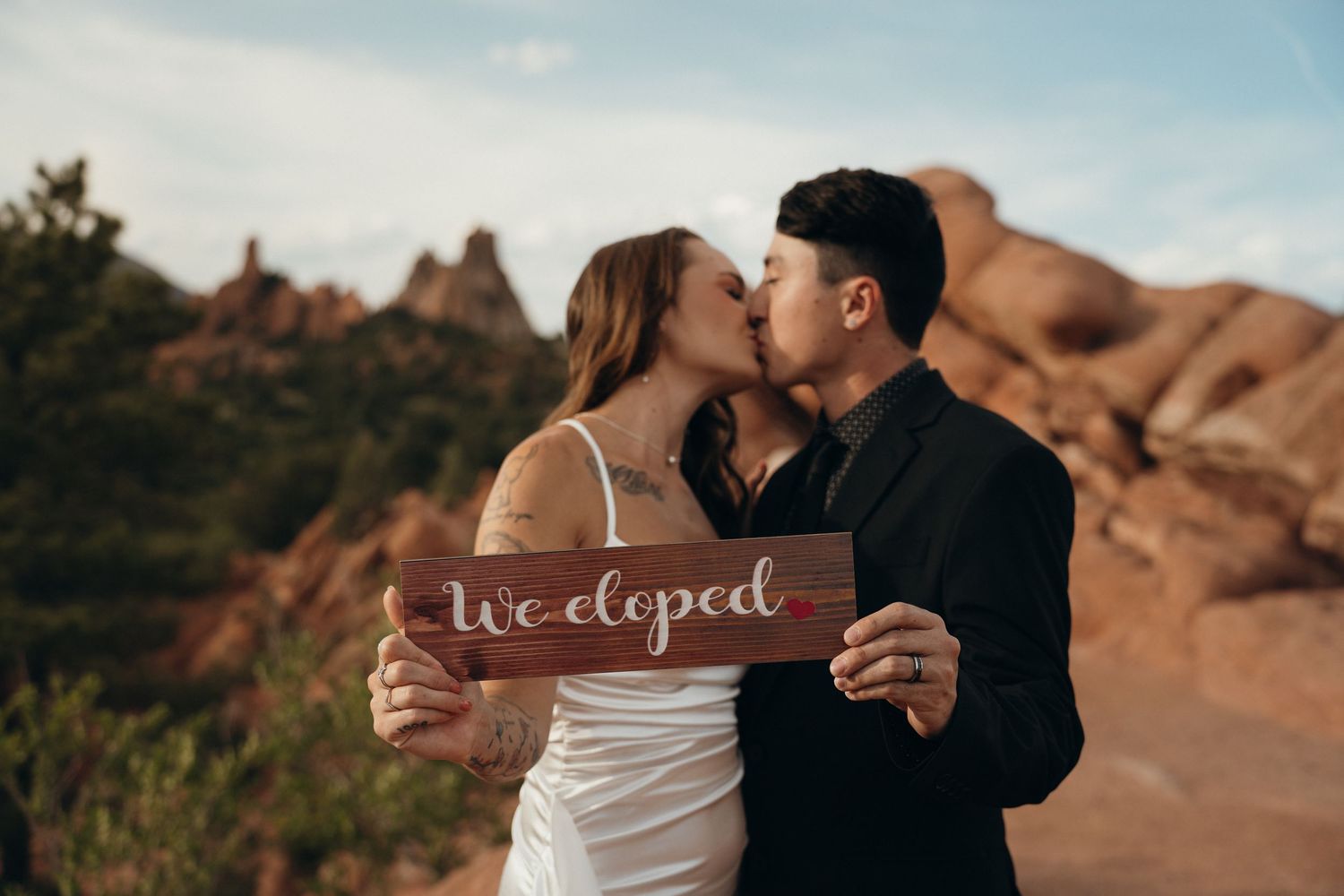 A couple kisses while holding a wooden 'We eloped' sign against a backdrop of red rock formations at sunset.
