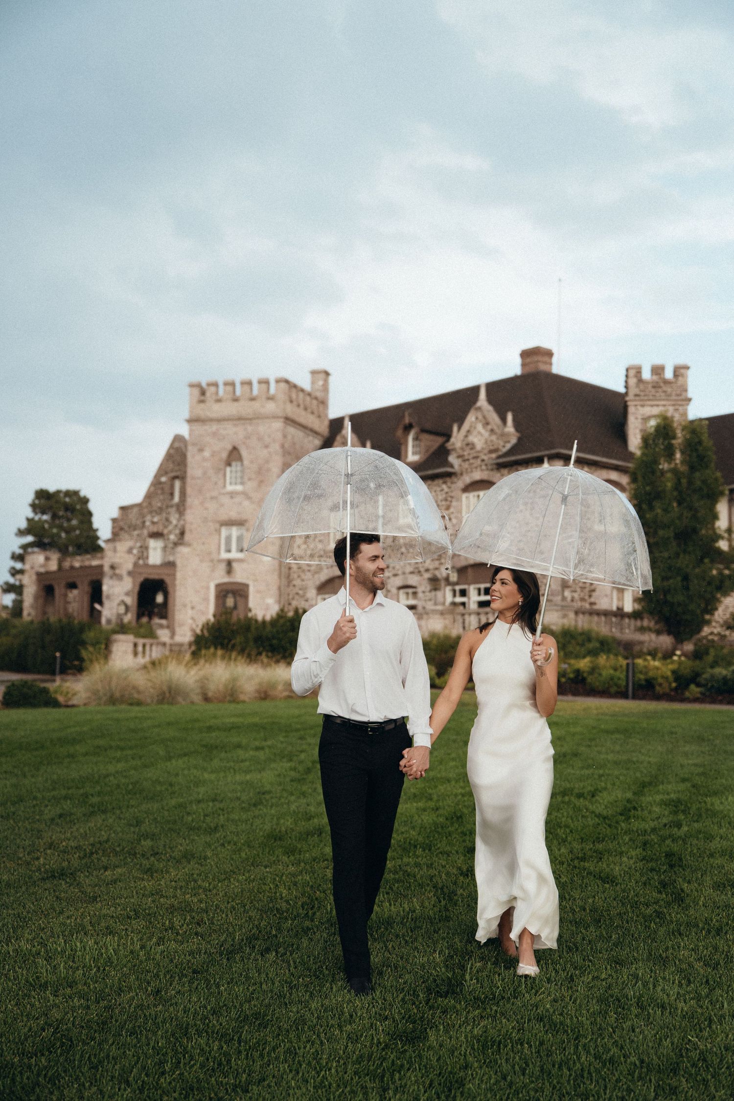 Couple in white attire walking with clear dome umbrellas on green lawn before a castle.