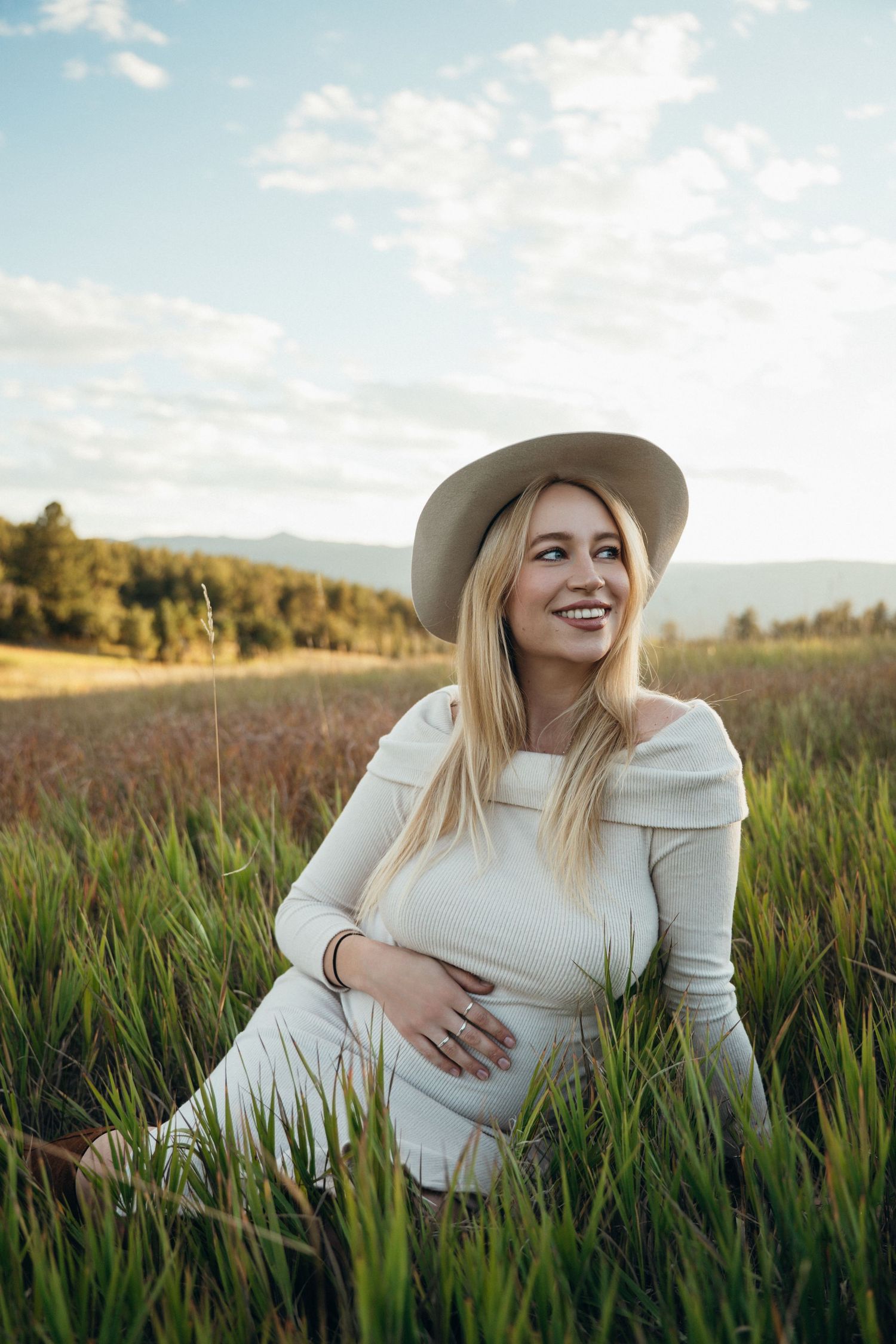 Woman in white sweater and hat sits in tall grass field at sunset smiling and enjoying nature.