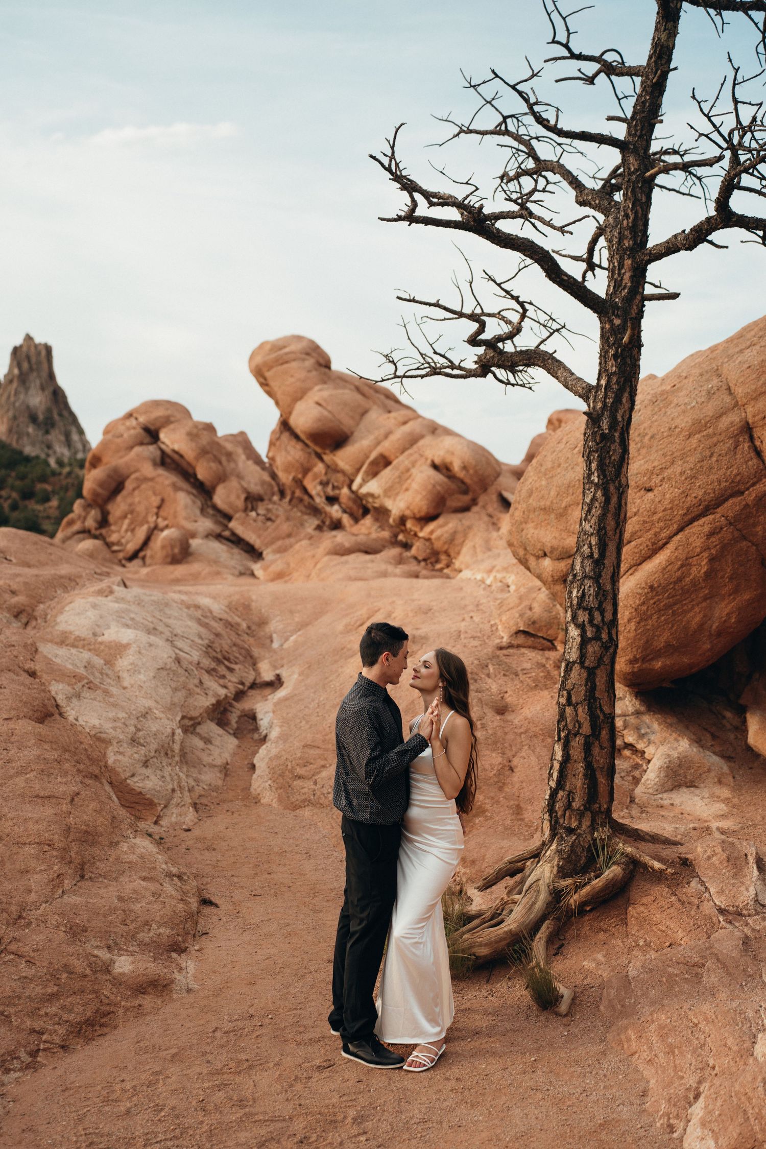 A romantic couple poses near a weathered tree against dramatic red rock formations in the desert.