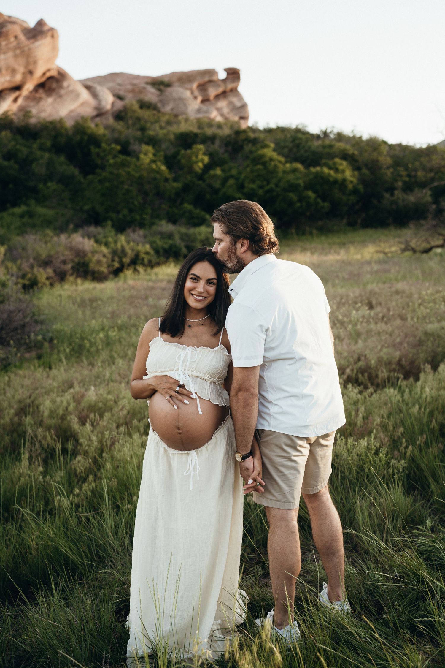 A couple in white outfits poses together for intimate maternity photos in a scenic grassy field at sunset.