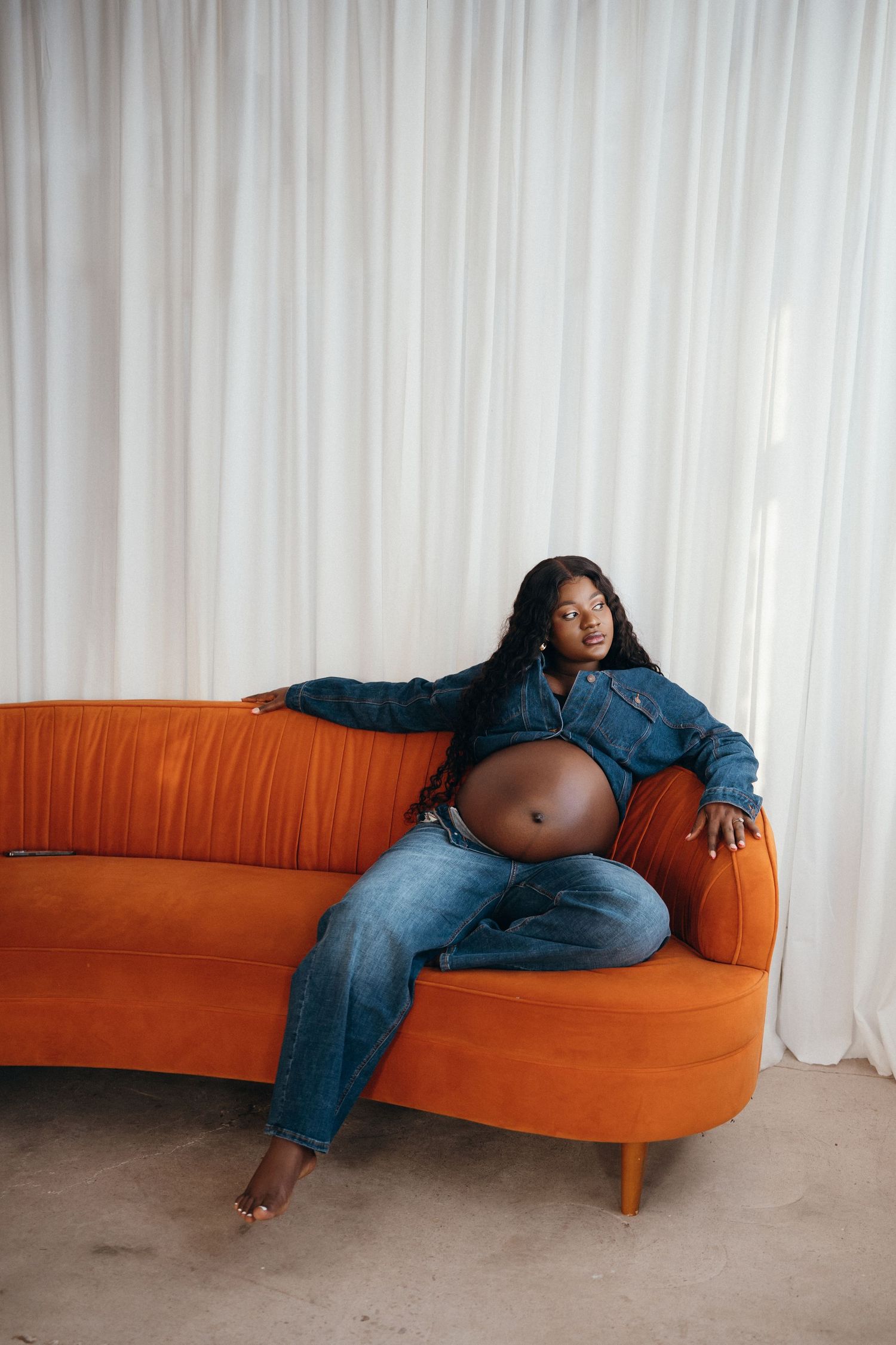 Person in denim outfit lounges on orange chaise lounge sofa in minimalist setting with white curtains.