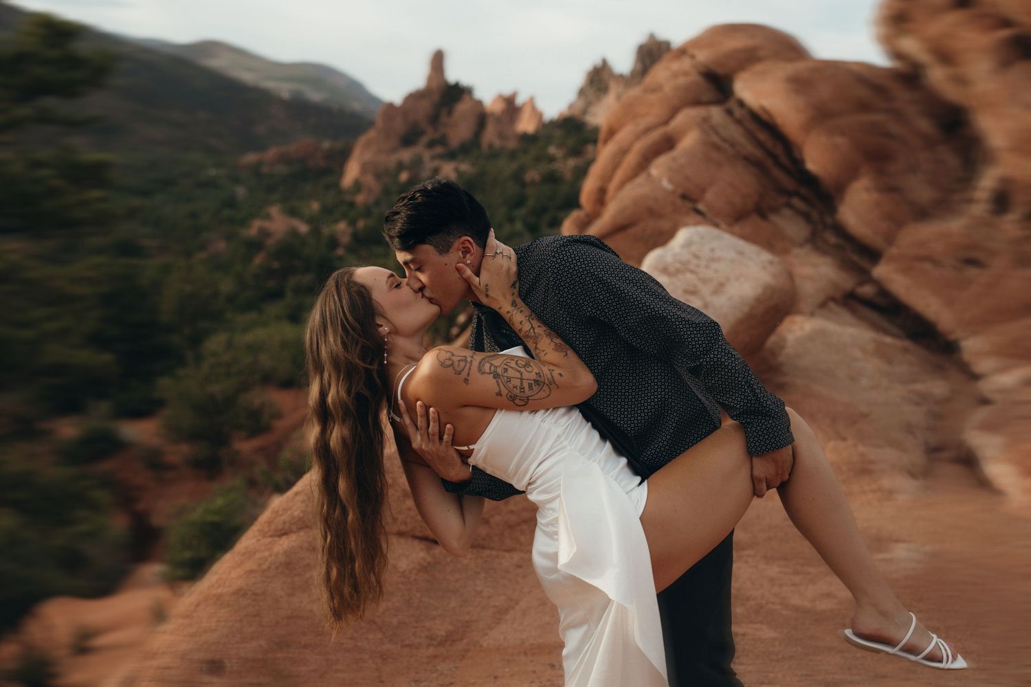 Romantic couple in formal attire dips and kisses at sunset against scenic red rock cliffs in desert landscape.