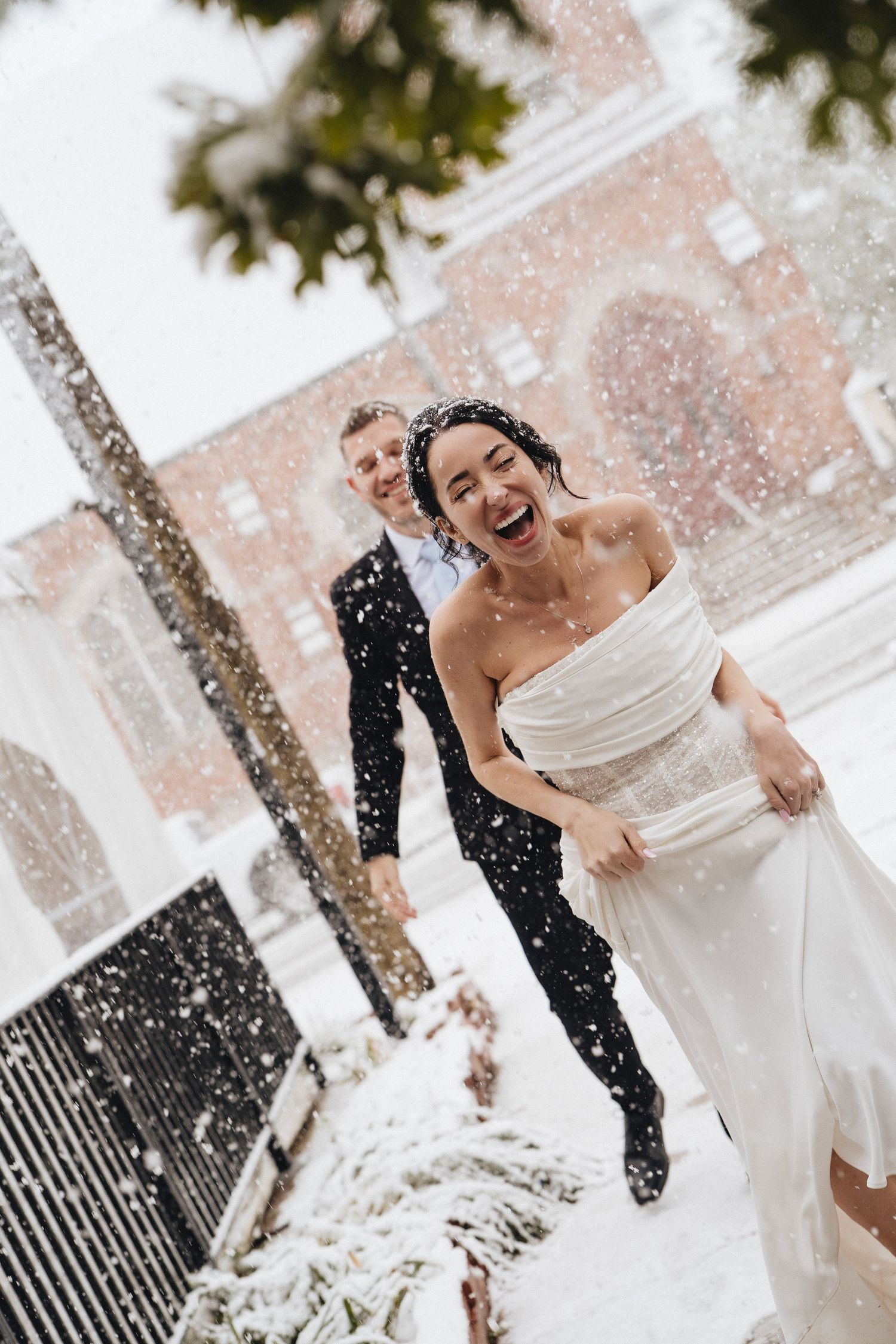 Joyful bride in white gown laughs in falling snow with groom in dark suit behind her, winter wedding outdoors.