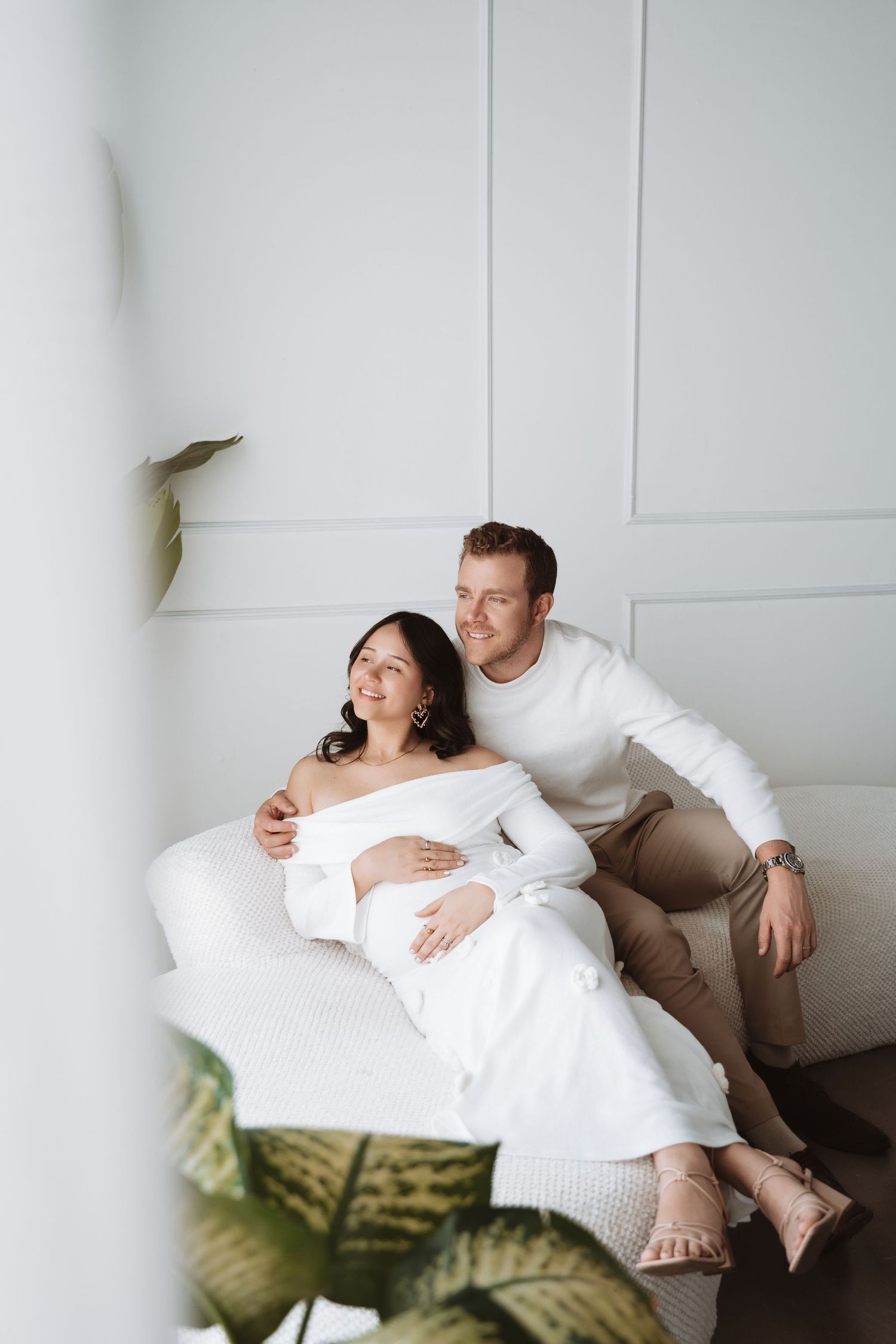 A couple relaxes together on a white couch in a minimalist room with a plant in the foreground.