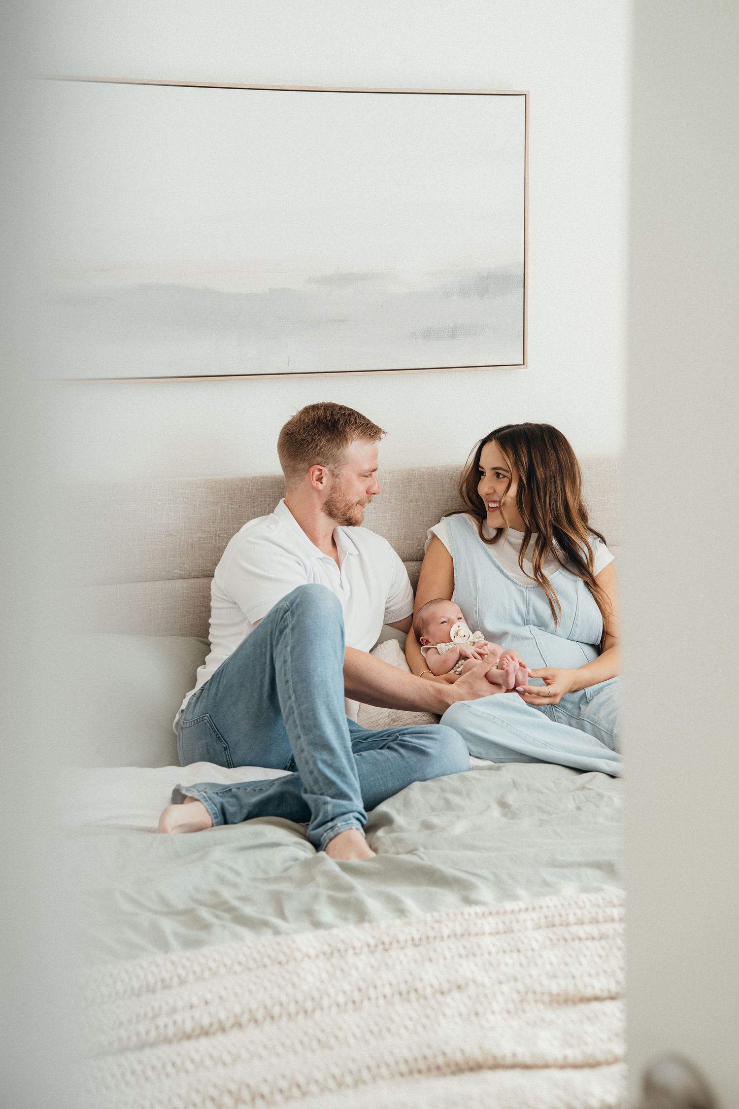 A couple sits on a bed together holding their newborn baby in a bright, minimalist bedroom setting.