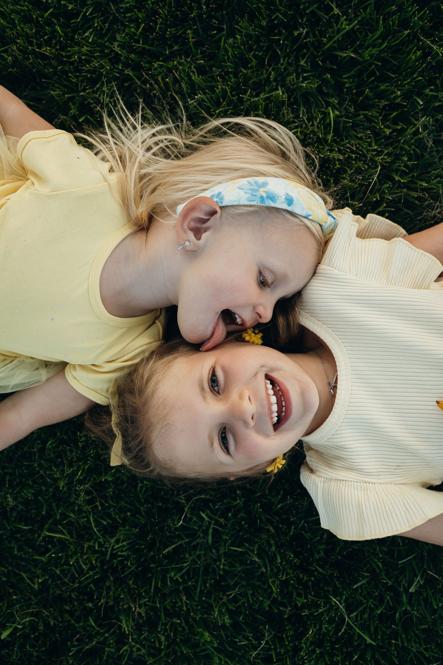 Two children lying on green grass head to head wearing yellow and white tops, smiling joyfully outdoors.