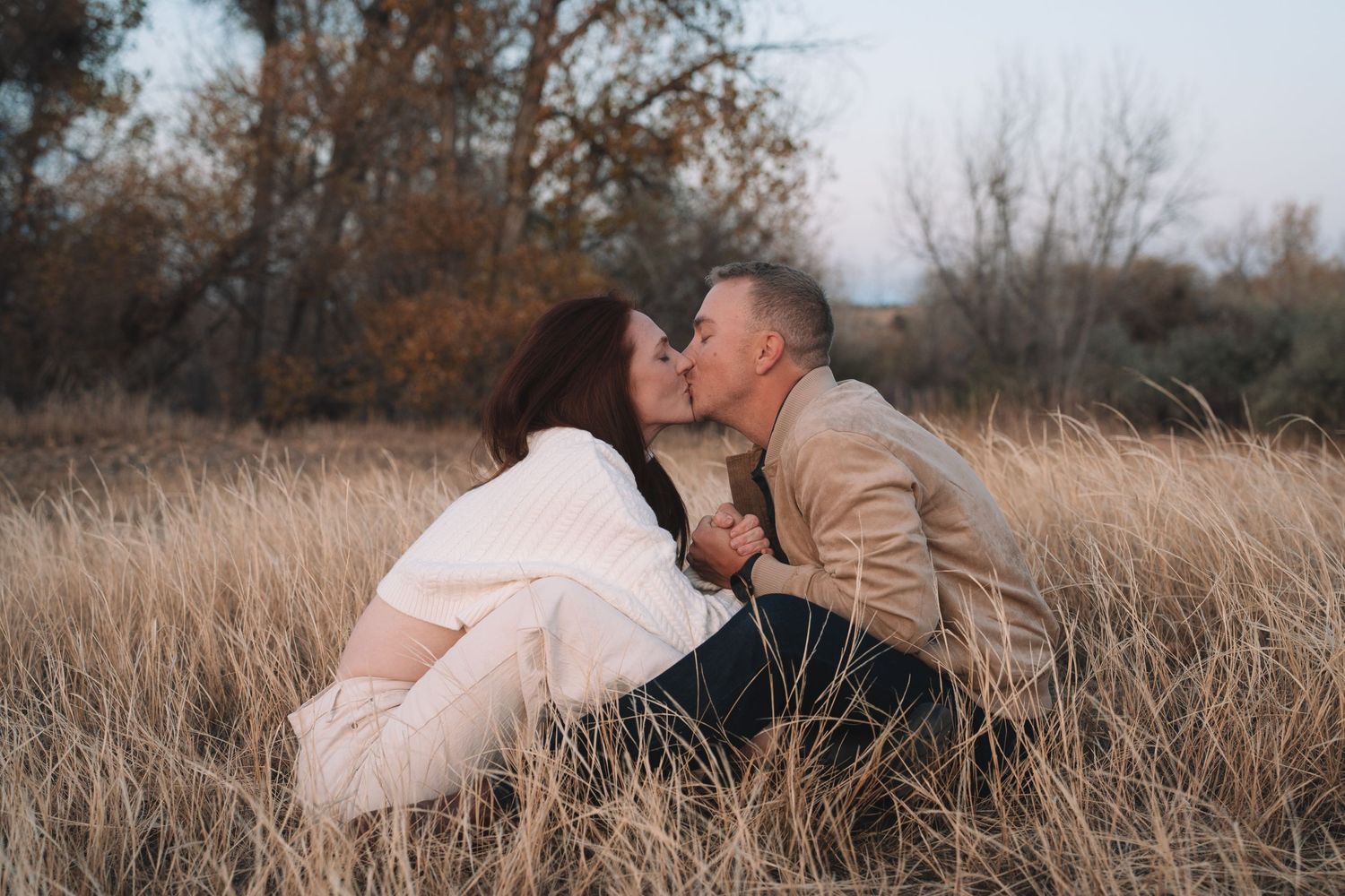 A couple shares an intimate moment while sitting in tall grass at dusk.