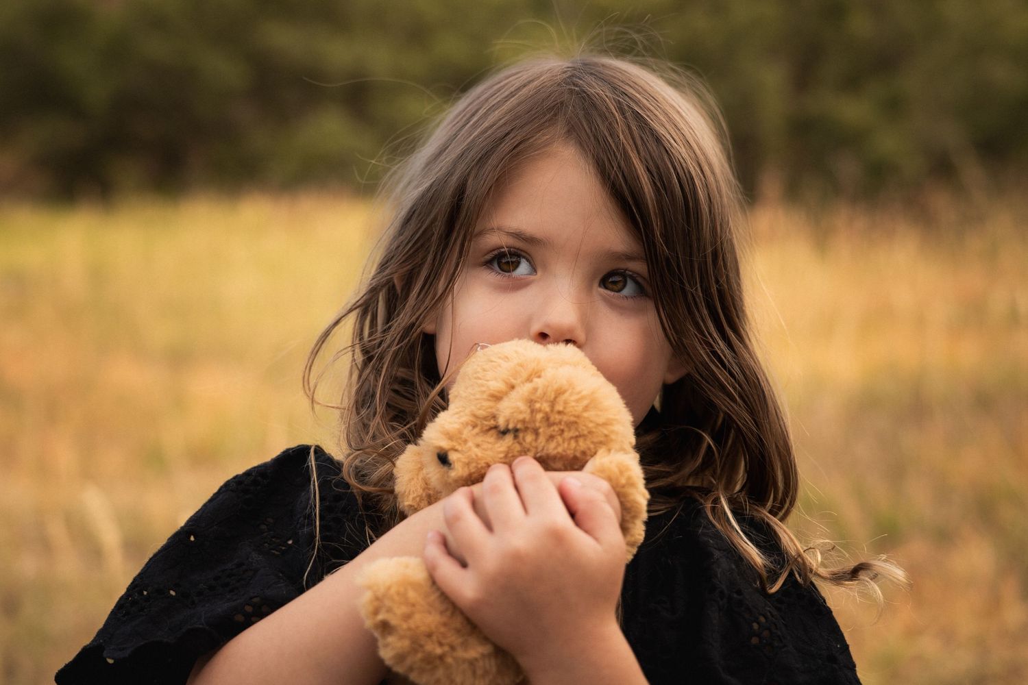 Child in black clothing holds brown teddy bear close to face in outdoor autumn setting.
