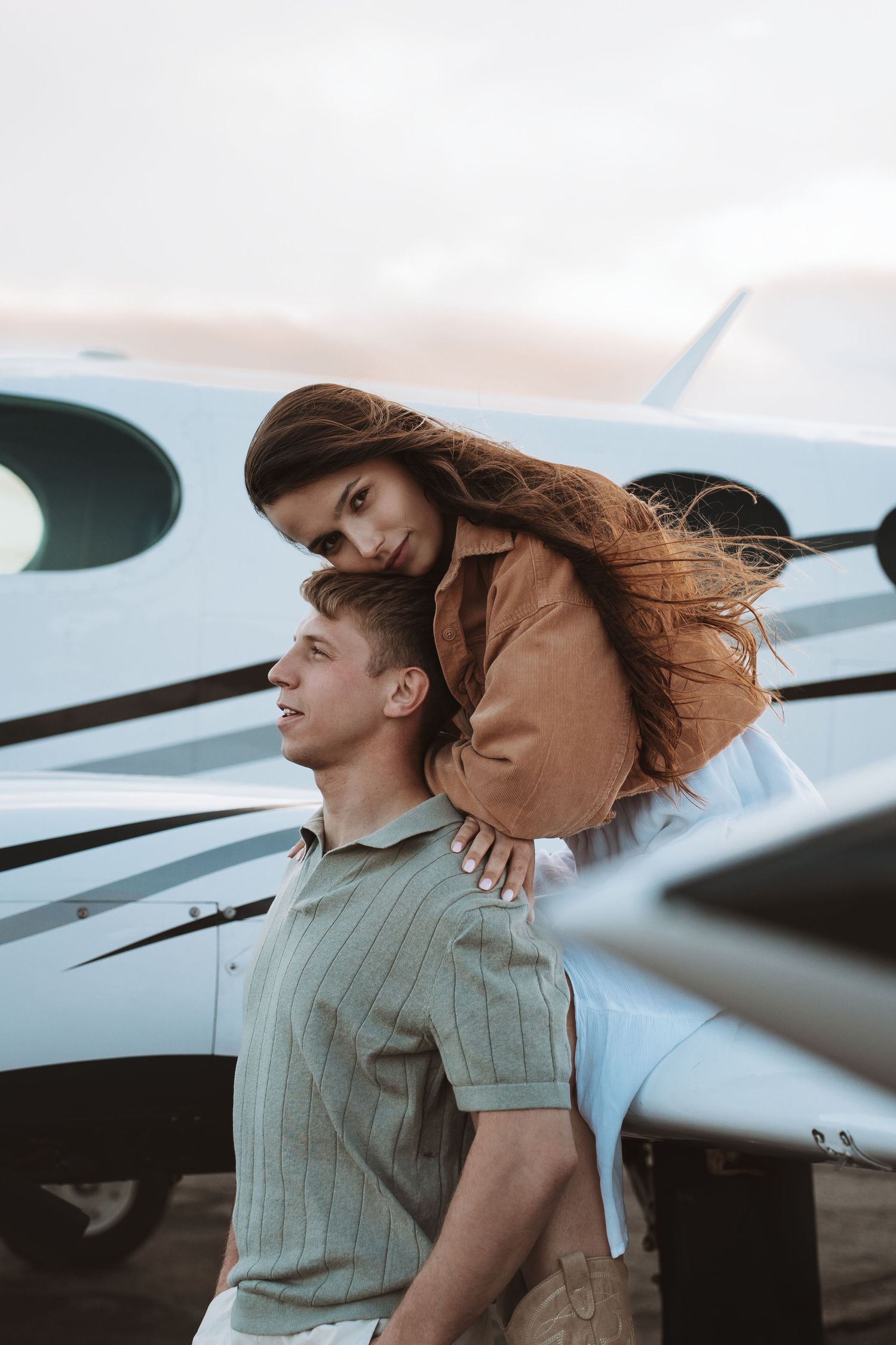 A romantic couple poses together near a small private airplane during sunset with her brown leather jacket flowing in the wind.
