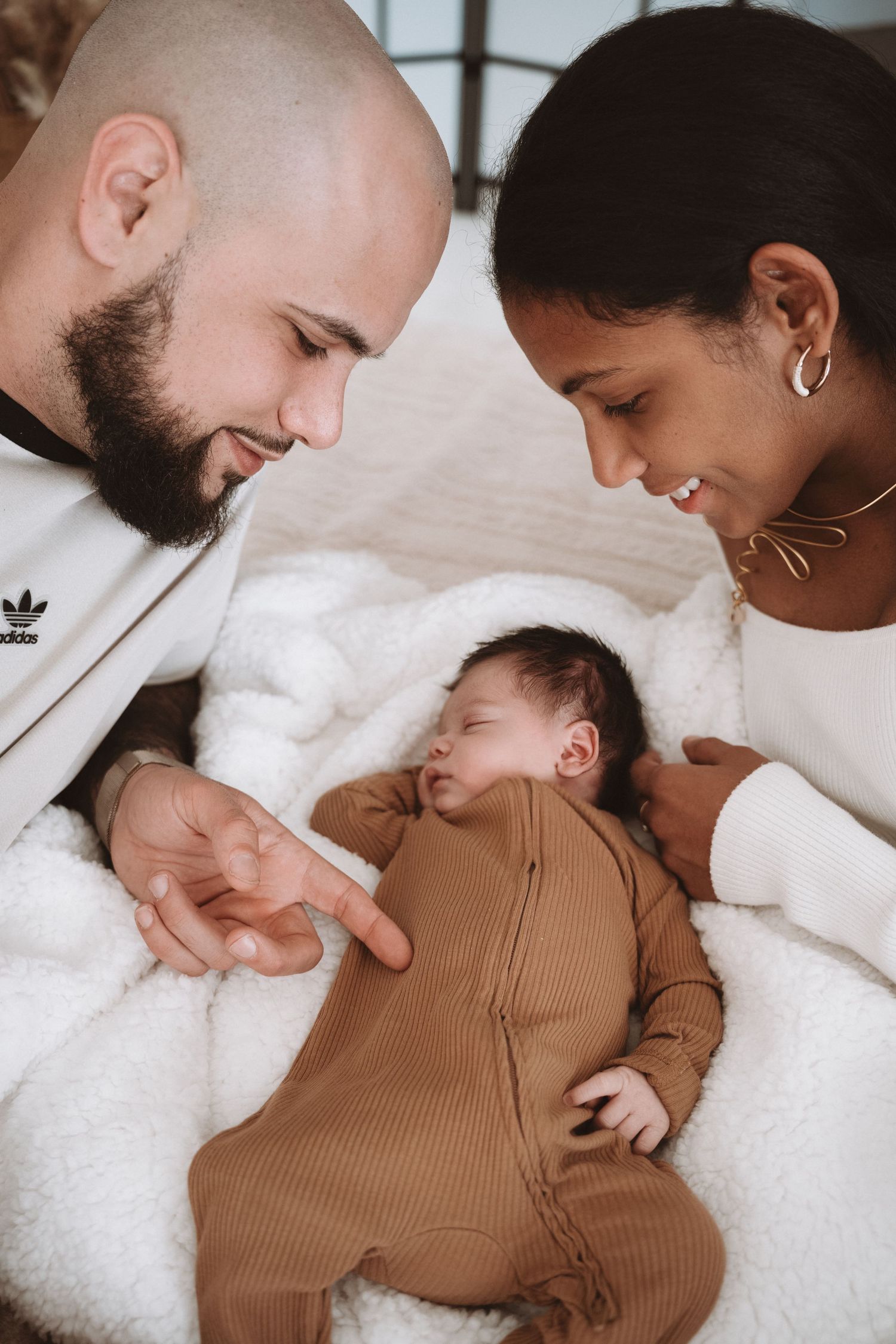 Parents lovingly watch over their sleeping baby in a brown ribbed onesie while lying on soft white blanket.