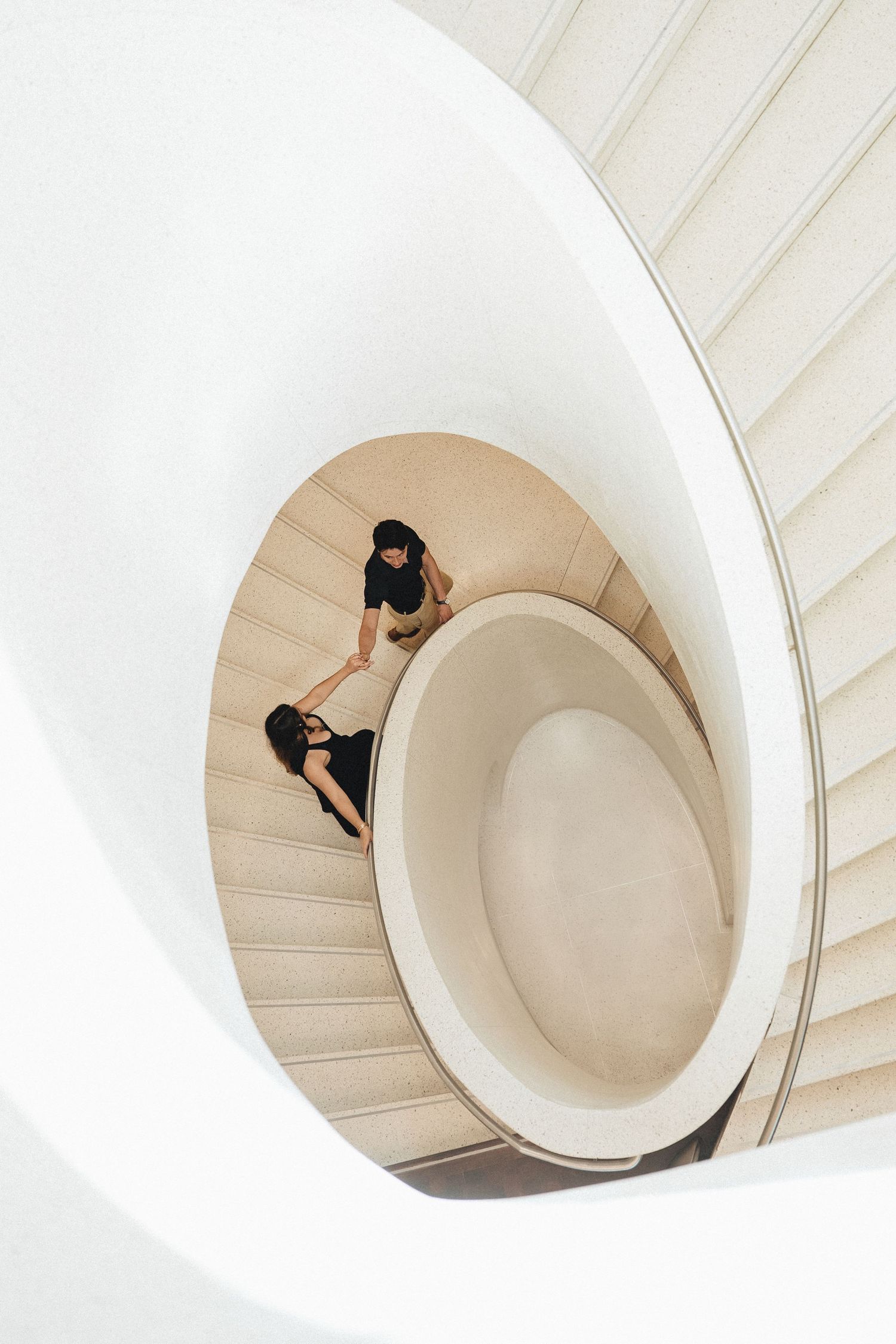 Two people in black outfits descend a stunning white spiral staircase, viewed from above in a modern architectural setting.