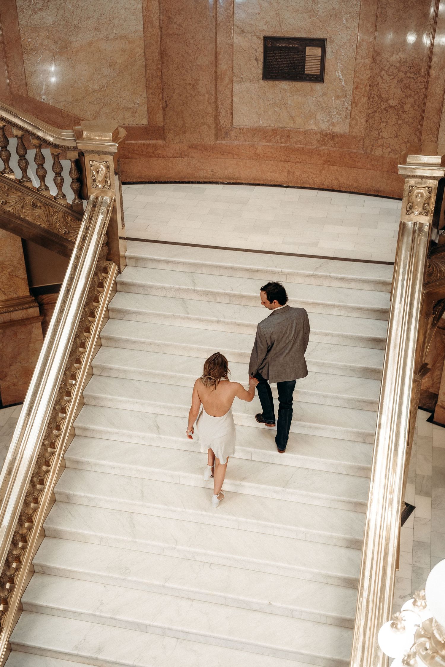 Two people ascending elegant marble stairs with ornate gold railings in a luxurious building interior.