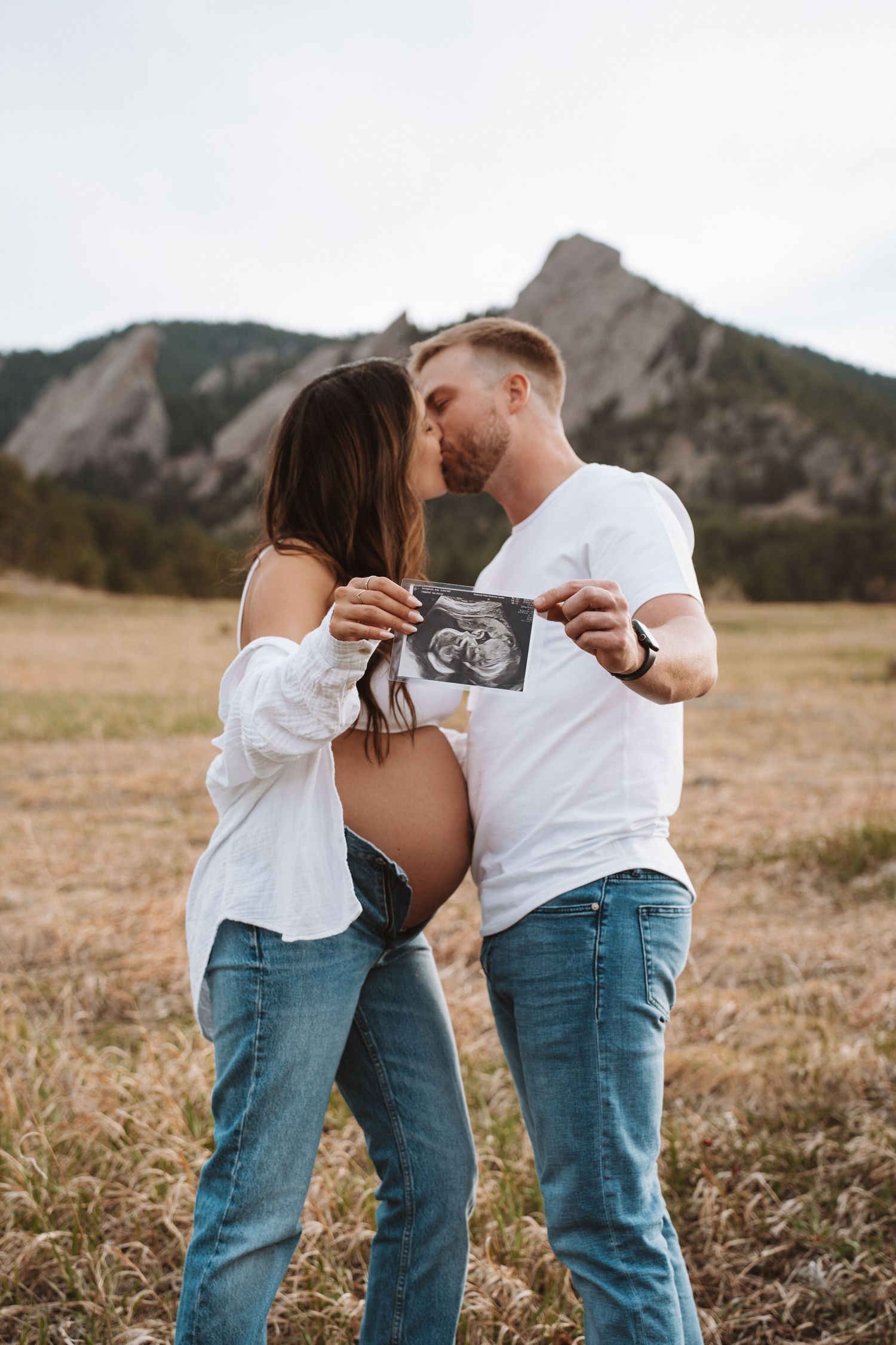 A couple shares an intimate moment holding an ultrasound photo in a field with mountains in the background.