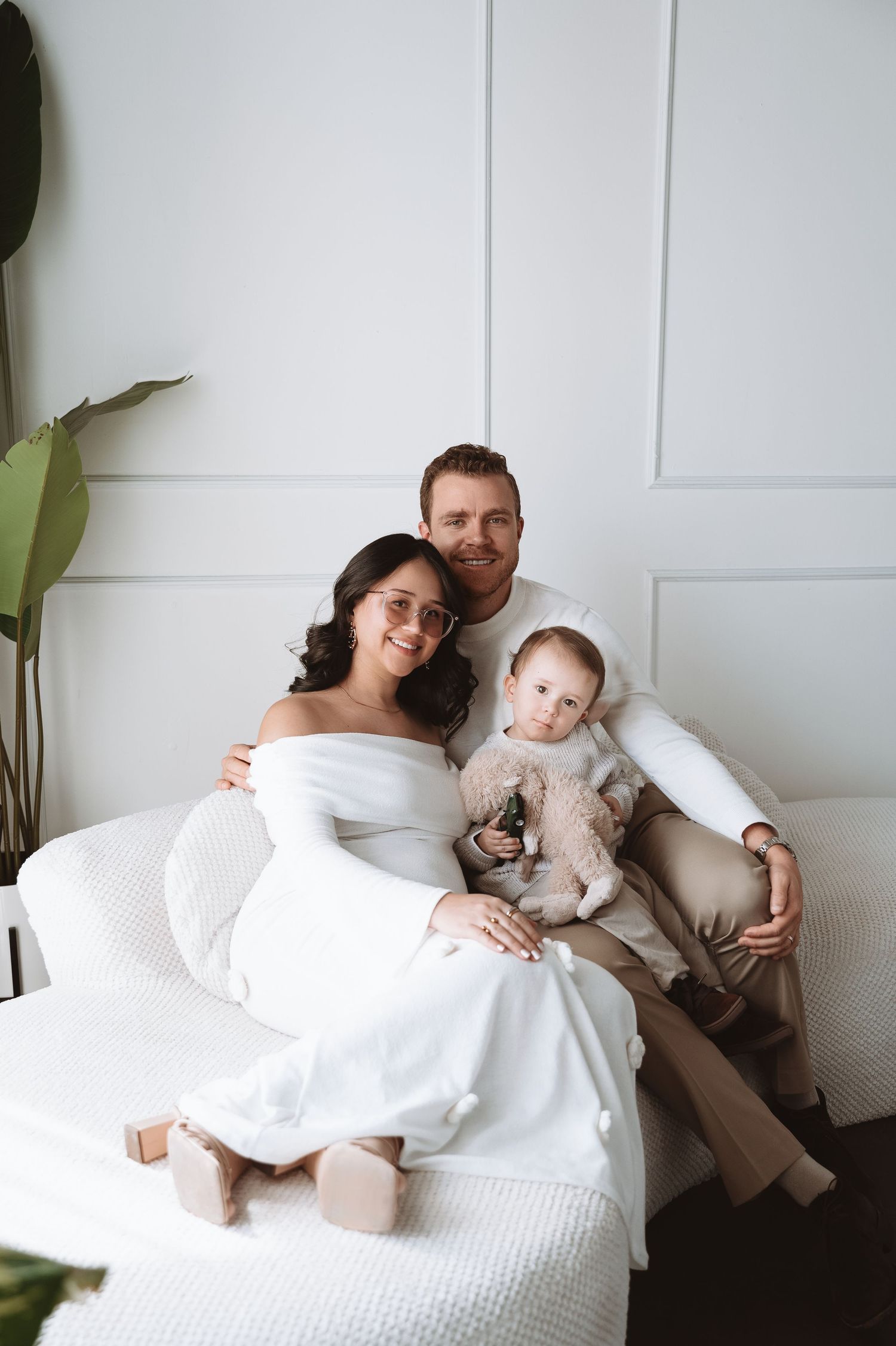 Family in all white clothing relaxes together on a couch with their small cat in a bright minimalist setting.