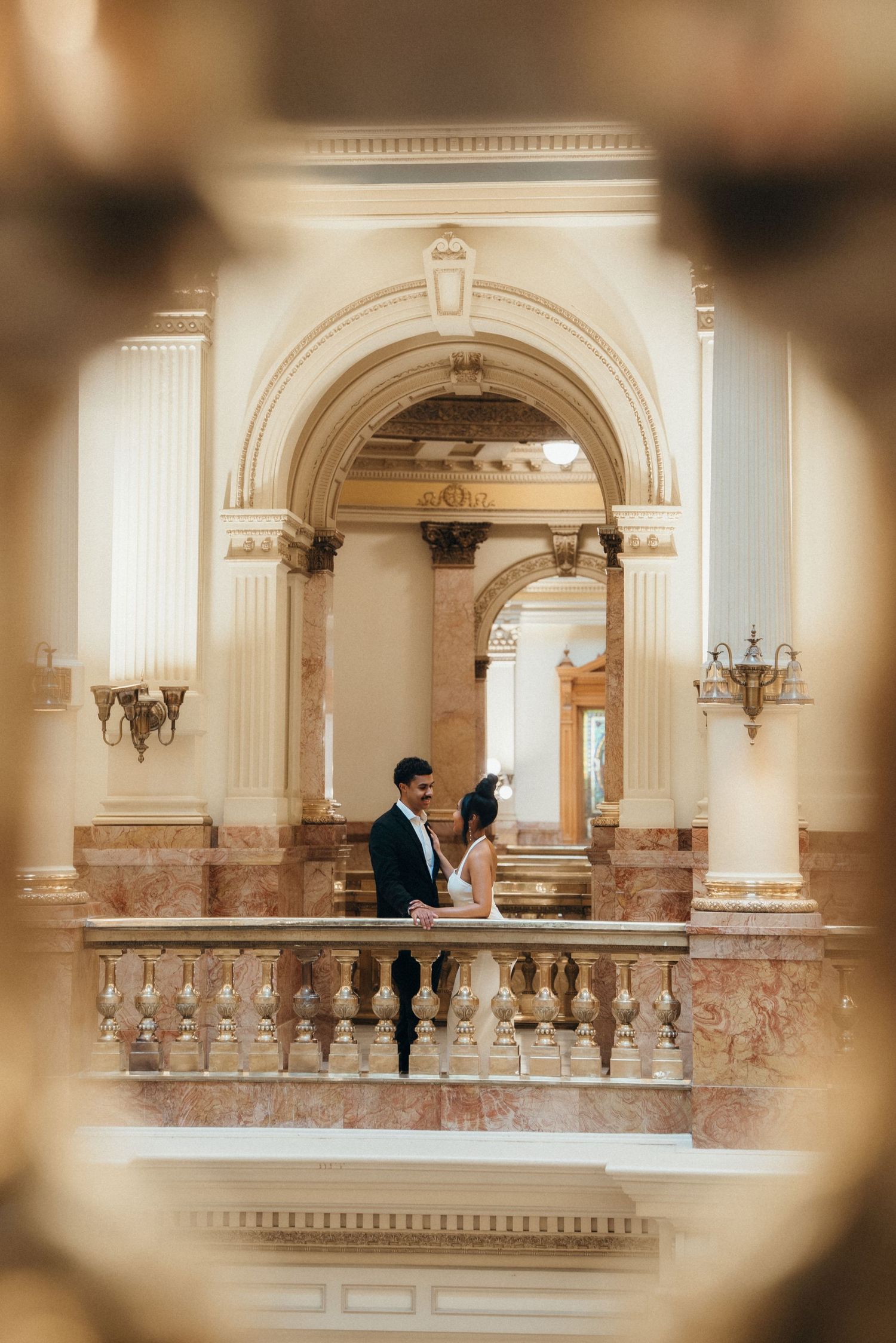Elegant sepia-toned panoramic view of a couple in formal attire on a grand marble balcony with ornate architectural details.