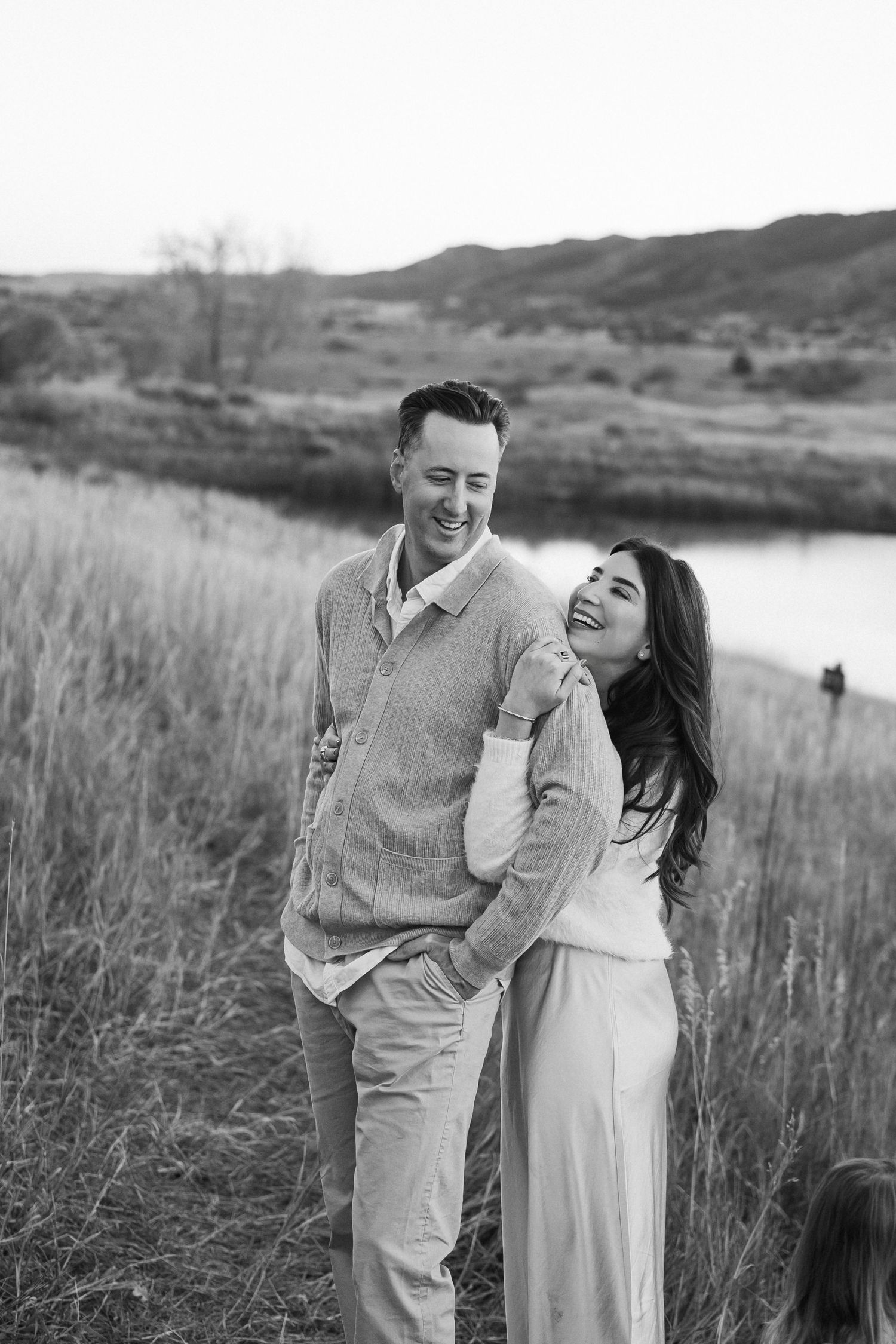 A couple shares joyful laughter together in a scenic mountain meadow with tall grass and rolling hills in black and white.