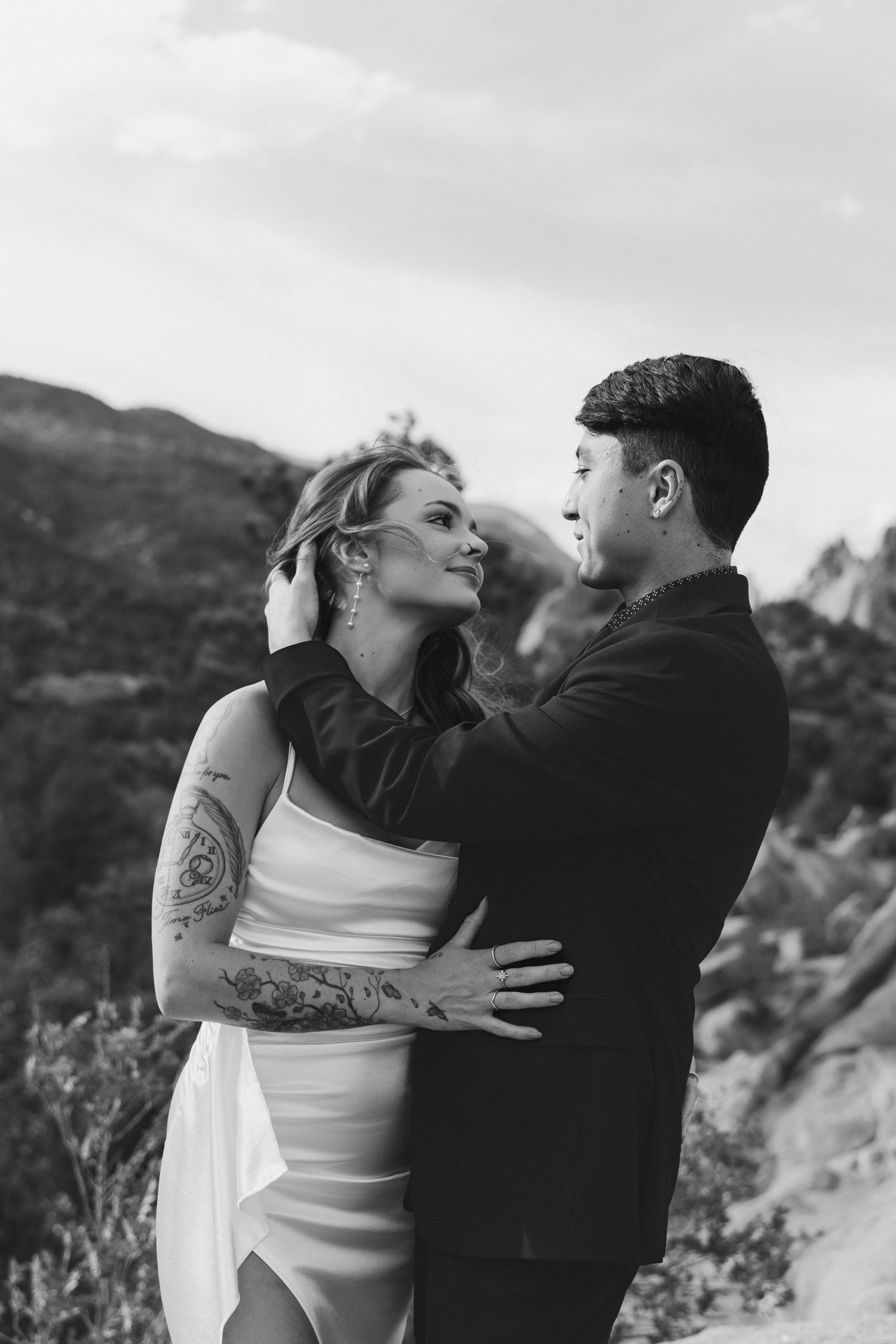 A romantic black and white portrait of a couple embracing outdoors with mountain peaks in the background.