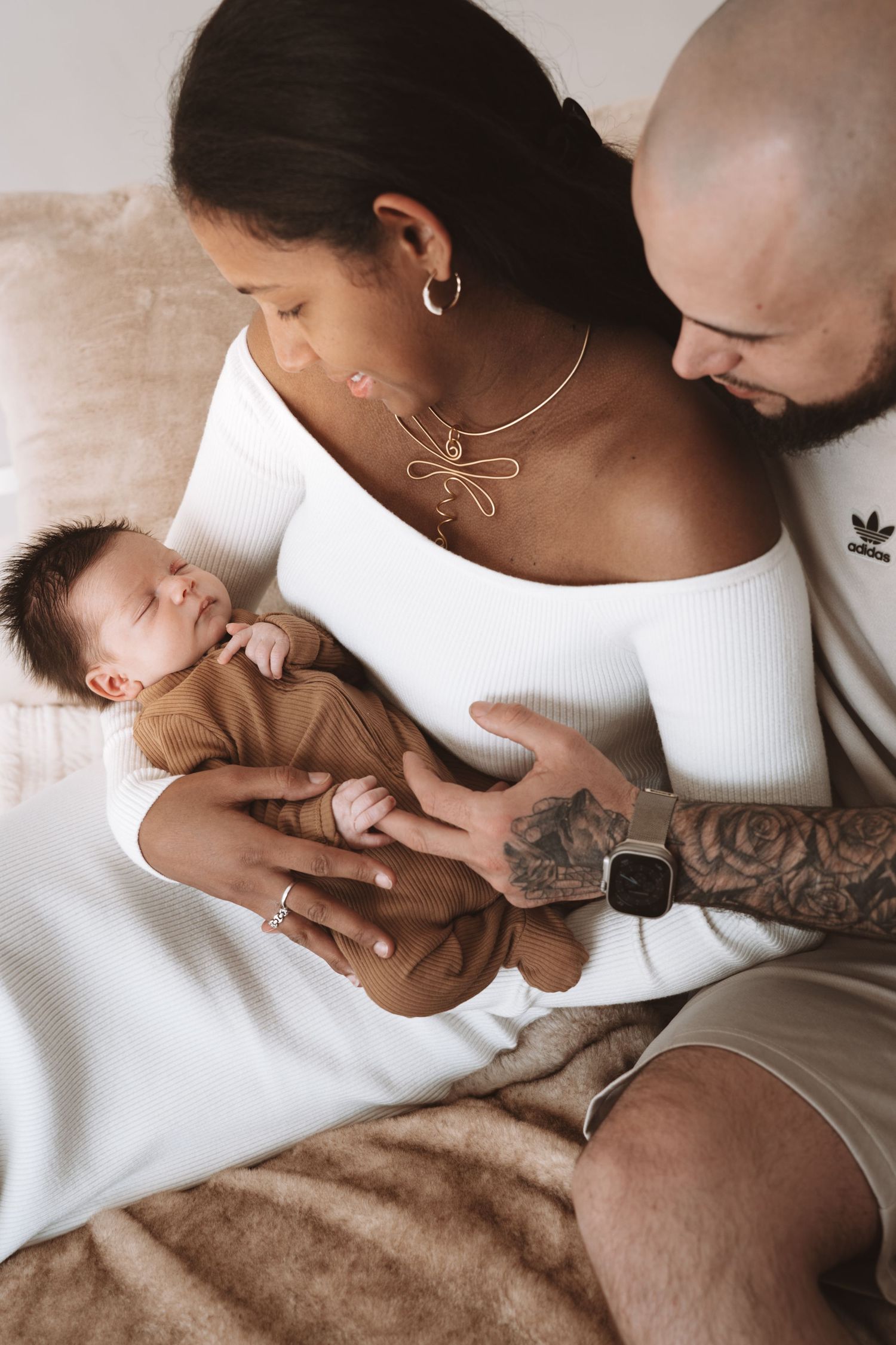 A tender family moment as parents gaze lovingly at their sleeping newborn baby wearing brown clothes on soft bedding.