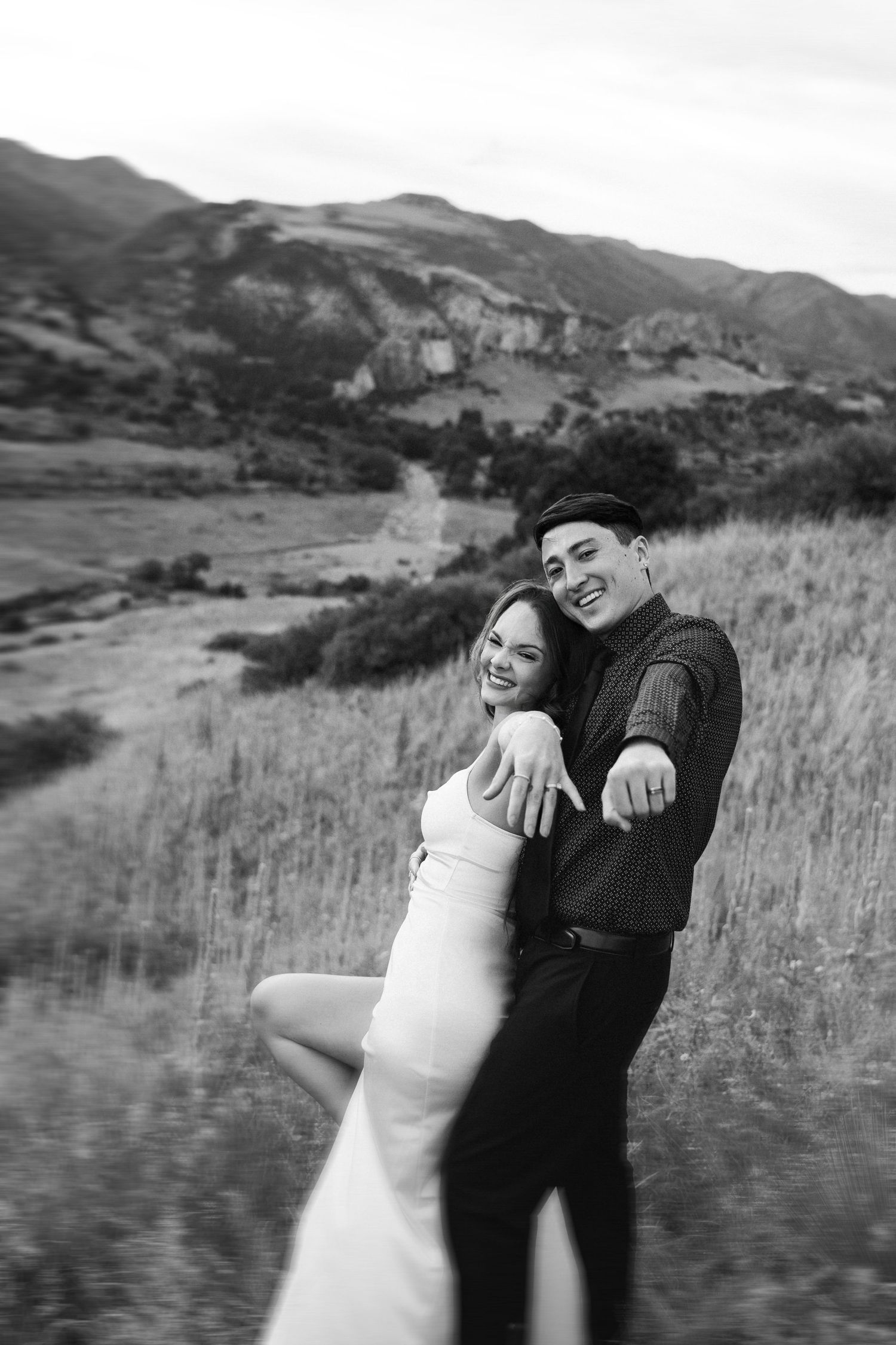 Black and white photo of a joyful couple posing together on a hillside with mountains in the background.