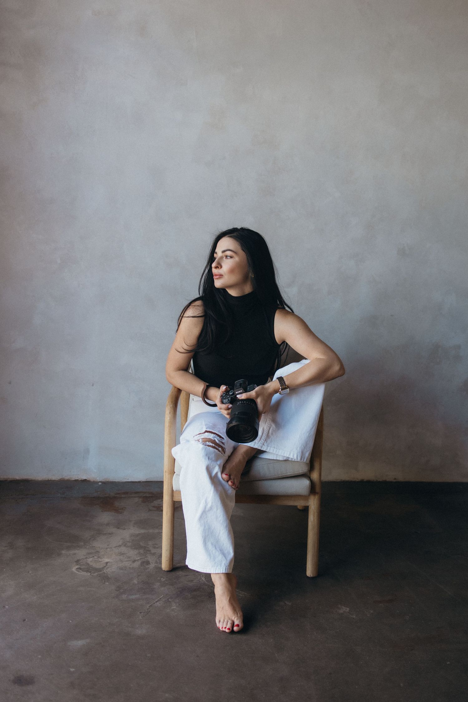 Woman in black top and white pants sits barefoot on wooden chair holding a camera, looking away thoughtfully.