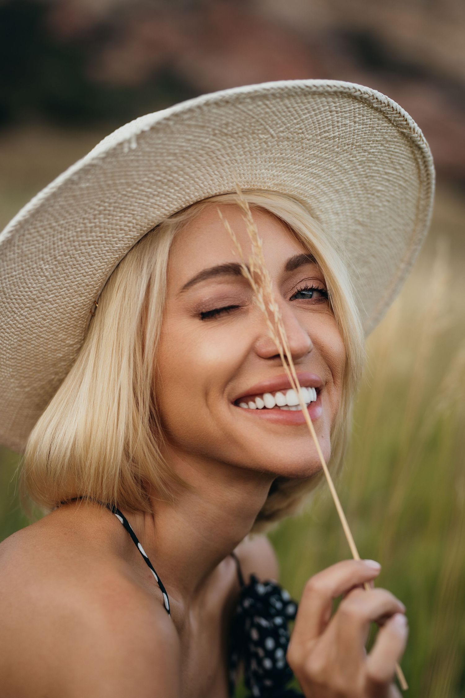 A person in a sun hat smiles while holding a dandelion flower in a summer field.
