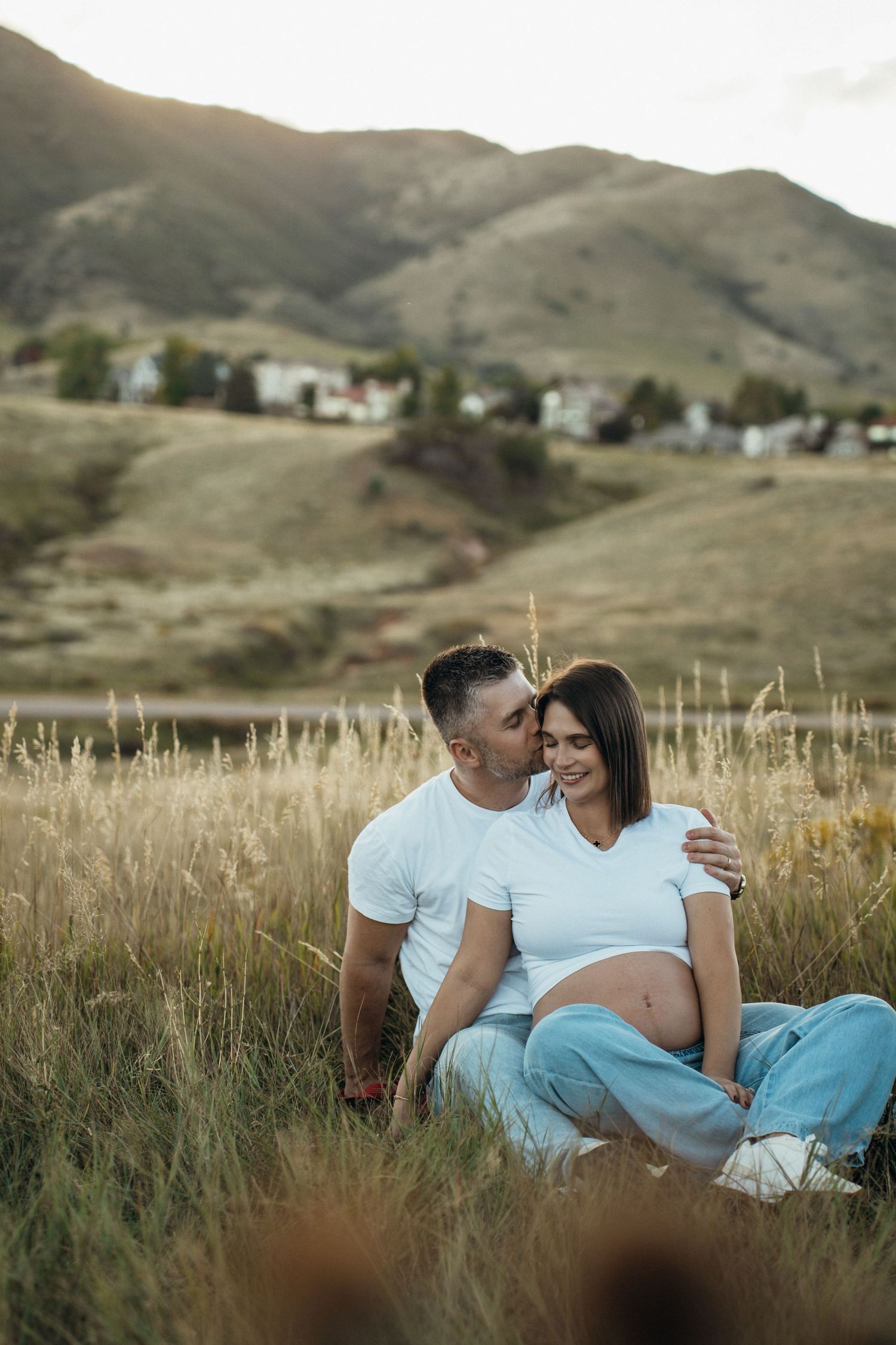 Couple shares intimate moment together while sitting in tall grass with mountains in background at sunset.