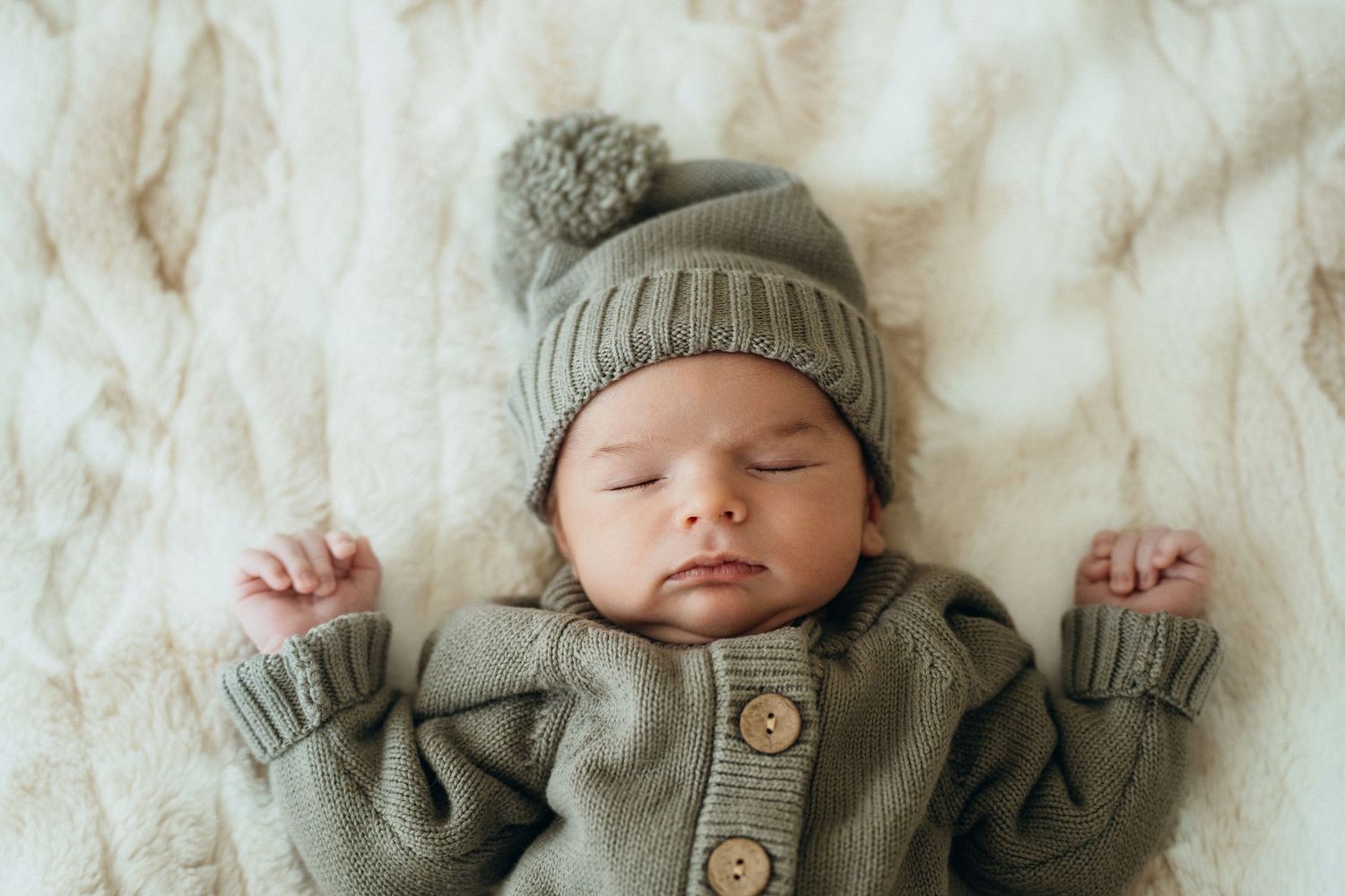 A sleeping baby wearing a cozy gray knit outfit and hat lies peacefully on white bedding.